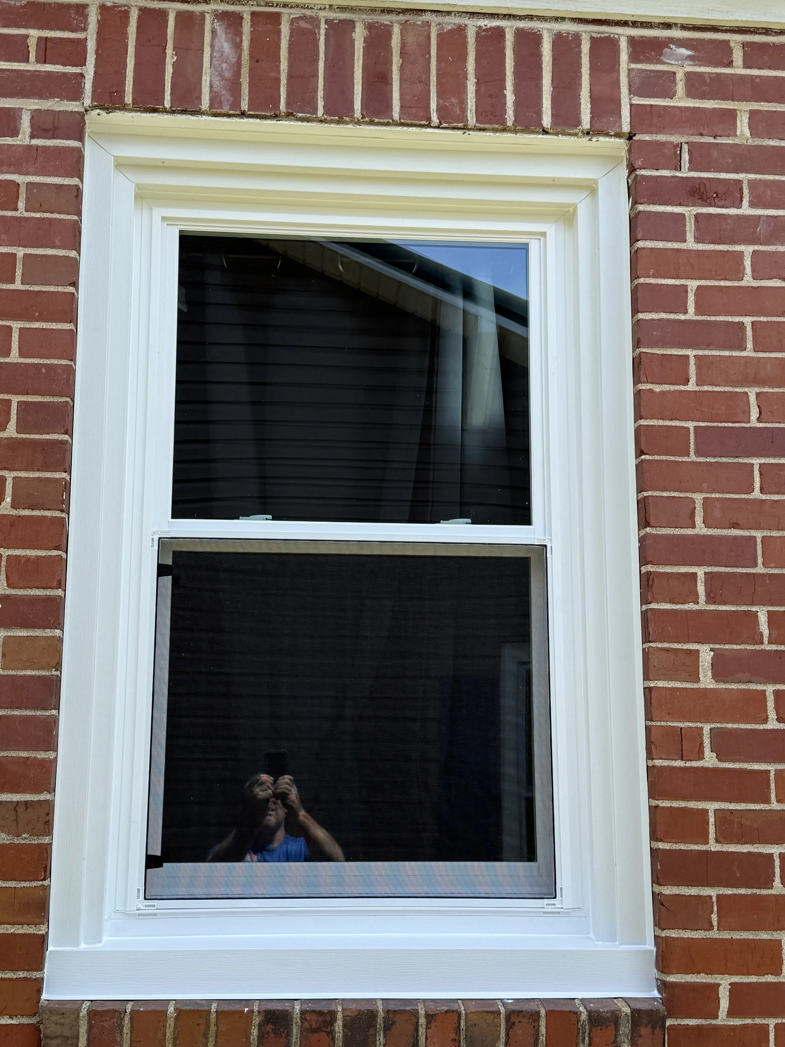 A window with a white frame set into a brick wall. The window has a screen, and behind it, black blinds are visible. There are reflections of the sky and a person taking the photo visible in the glass.