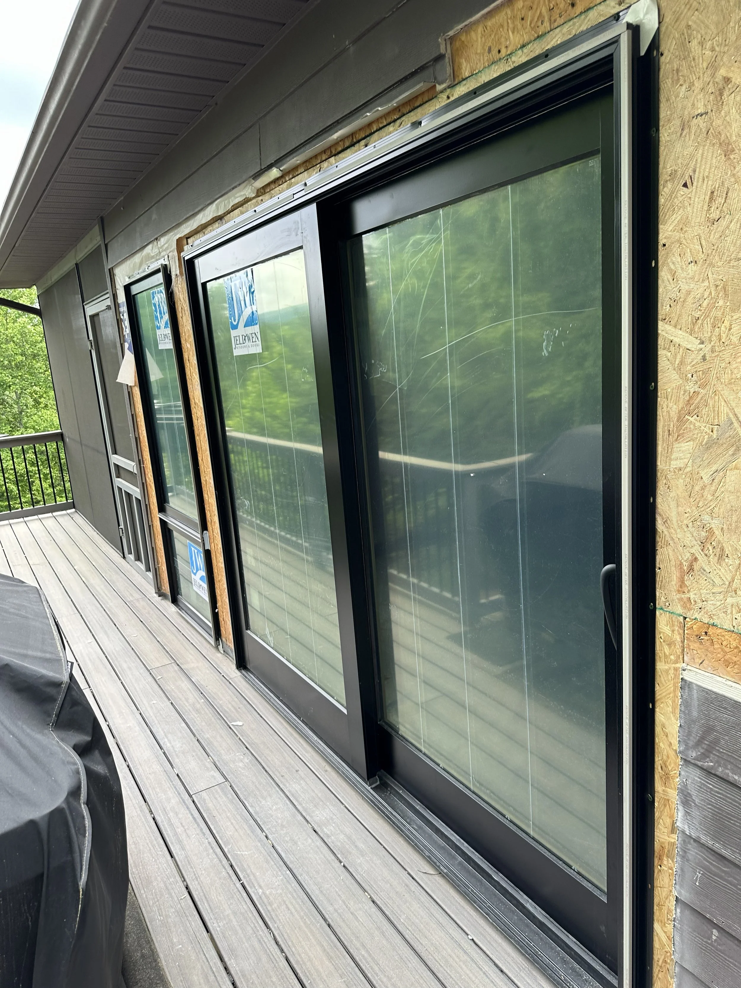 Two large black-framed glass sliding doors installed on a wooden wall exterior, with construction materials visible around them, on a wooden deck.