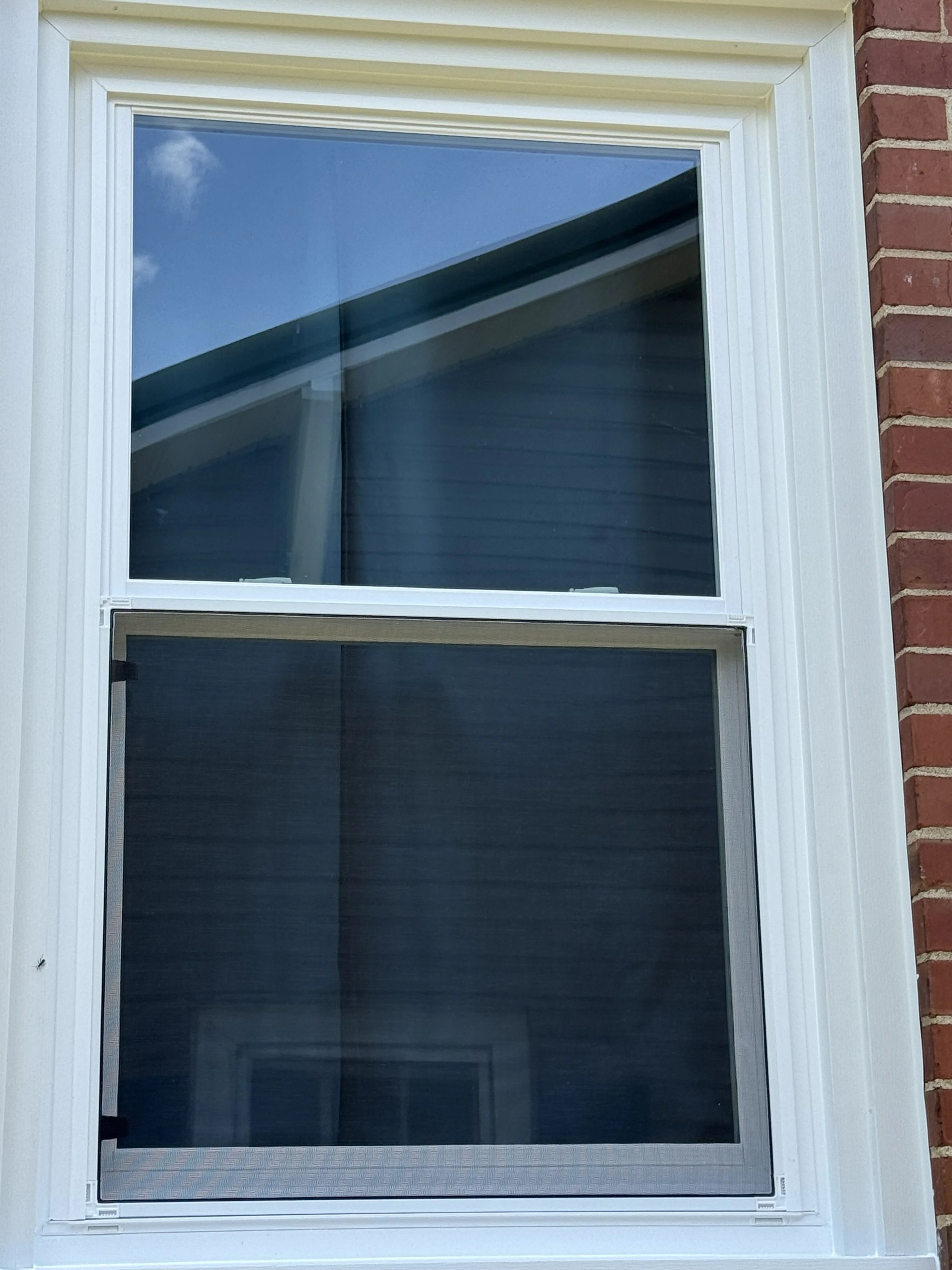 A close-up view of a white-framed window with a screen, set into a brick wall. The window's glass reflects a blue sky with a few clouds.