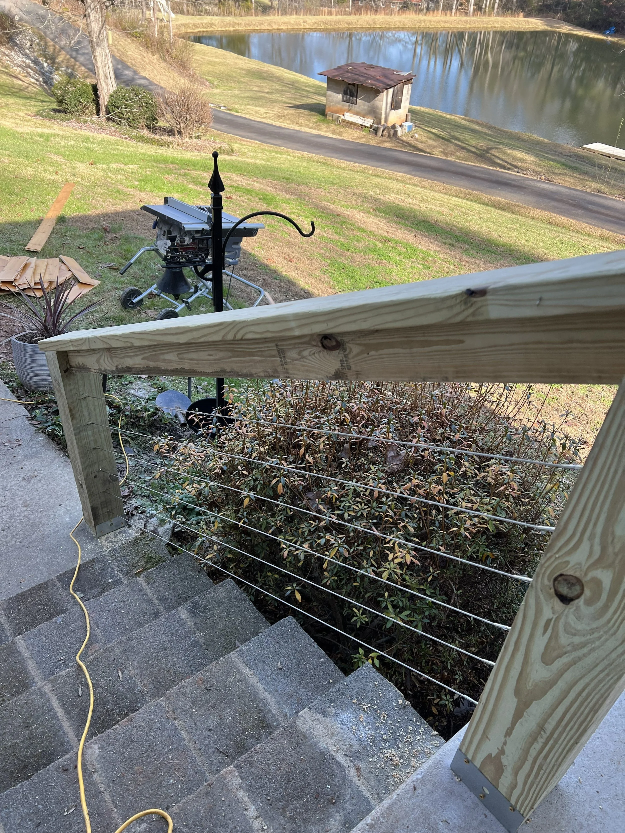 A backyard scene with a staircase going down to a grassy yard, a wooden railing, and a view of a small pond or lake surrounded by trees. There is some construction material and a small outside grill or stove on the patio area.