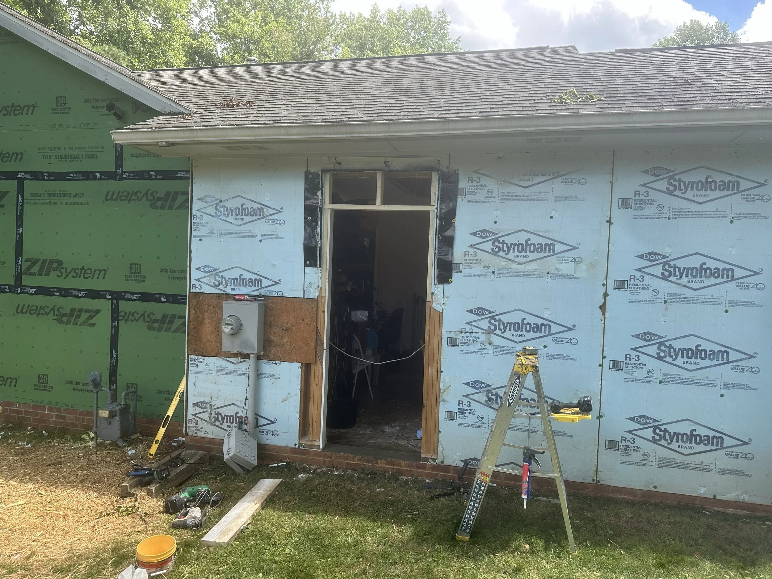 Under construction house with partial siding and opening for door, construction tools and materials on lawn, cloudy sky.