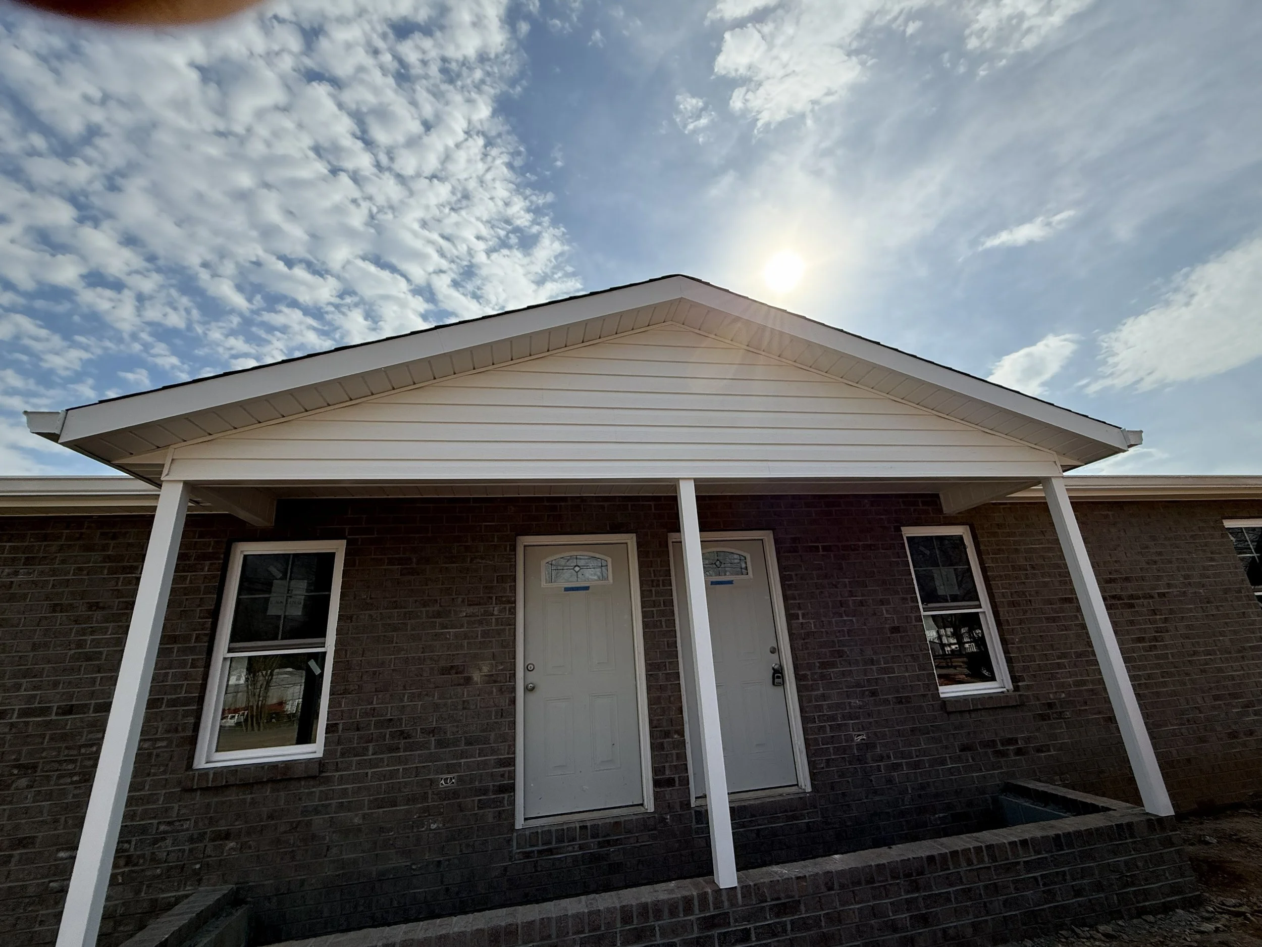 The photo shows the front of a house with a white door, a brick facade, two windows, and a porch with white columns. The sky above has scattered clouds and the sun is shining.
