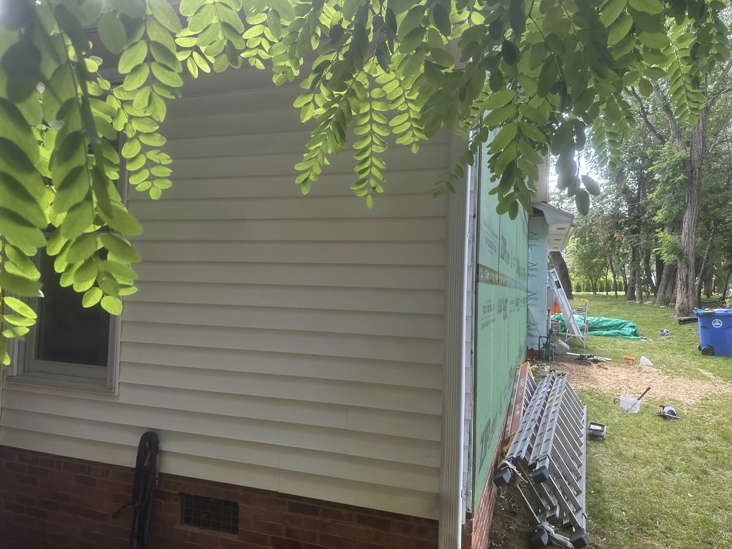 Close-up of a tree with green leaves in the foreground and a house under construction or renovation in the background. Building materials and tools are visible near the house.