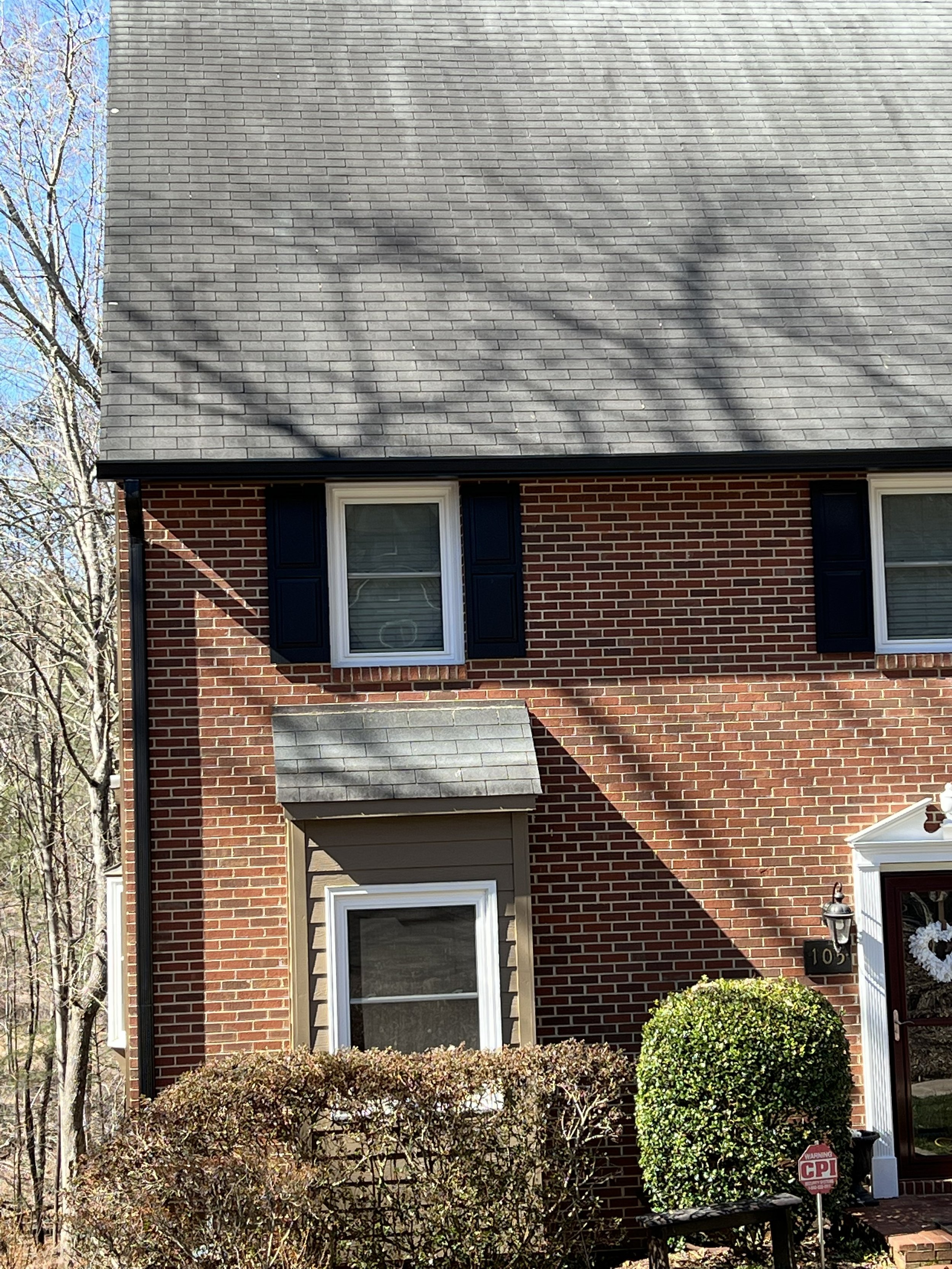 Front view of a brick house with black shutters and a small porch, surrounded by bushes and trees with shadows cast on the roof.