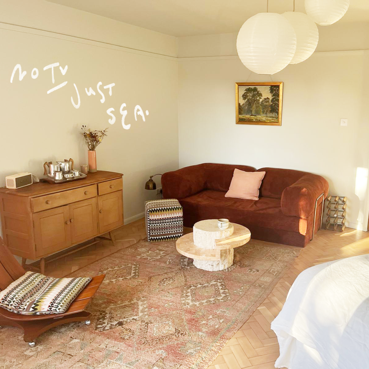 Living room with a brown sofa, a patterned armchair, a wooden sideboard with tea sets and a pink vase, a framed landscape painting, paper lantern ceiling lights, and a decorative rug.