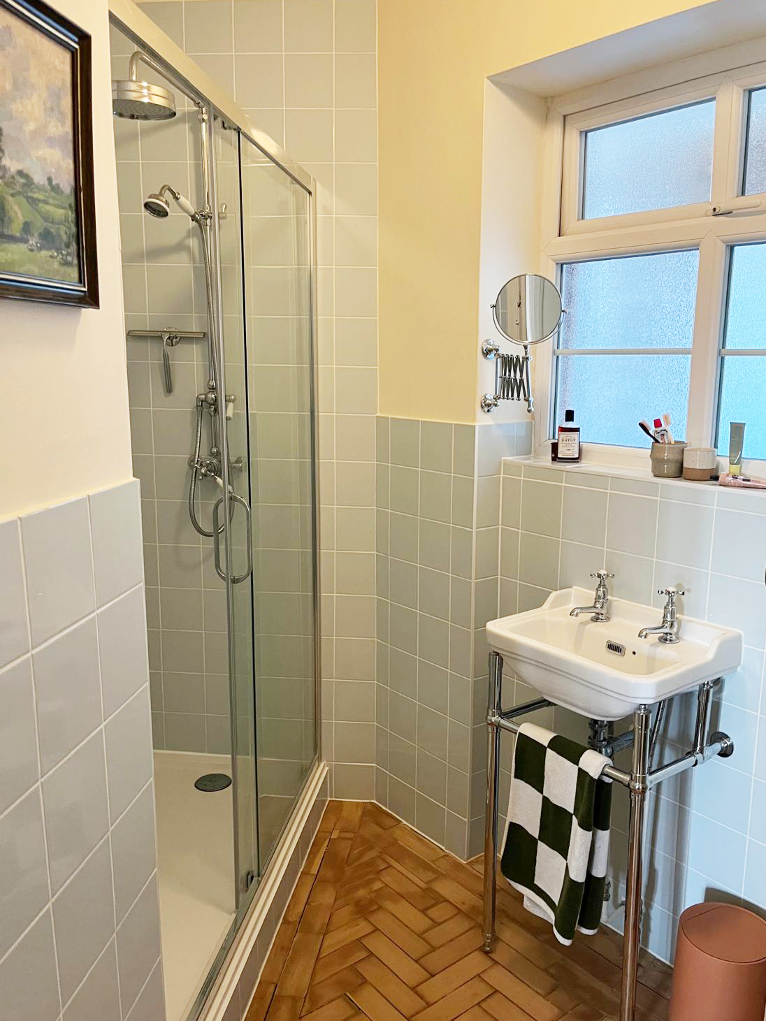 Small bathroom with a walk-in shower, a wall-mounted sink with a checkered towel, a window, and a small round mirror.