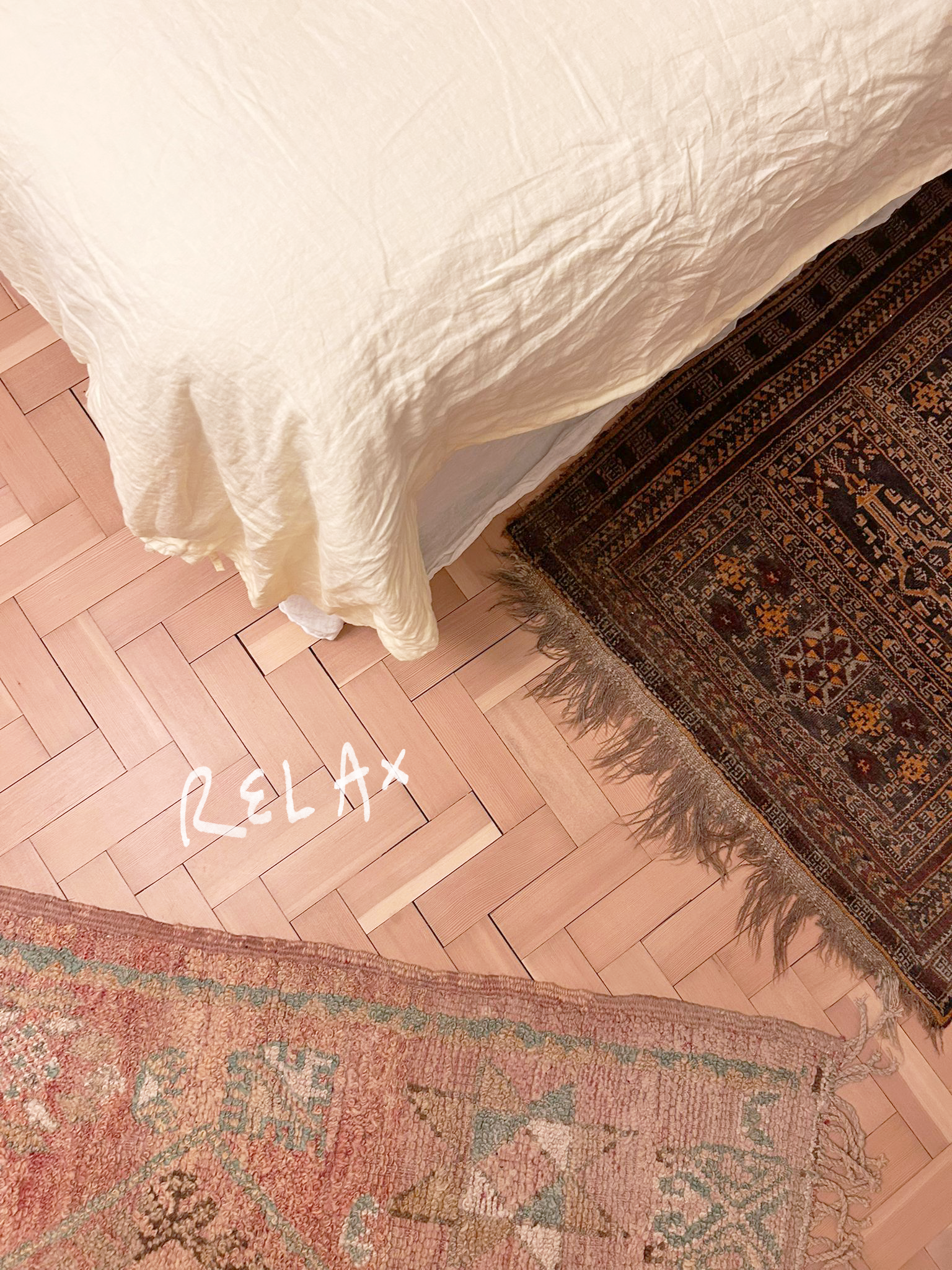 Close-up of a bed with a cream-colored blanket partially covering a dark, patterned rug on a wooden floor. The word 'RELAX' is handwritten in white near the bottom left corner.