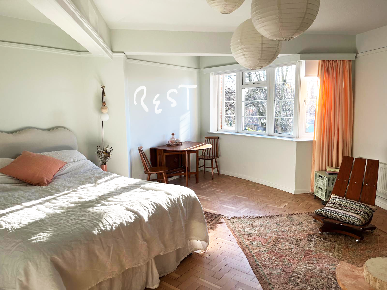 A bedroom with a large bed on the left, white bedding, a peach-colored pillow, and a small vase with dried flowers. Sunlight streams through a large window with orange curtains. A wooden table with two chairs is in the far corner, and a vintage chair