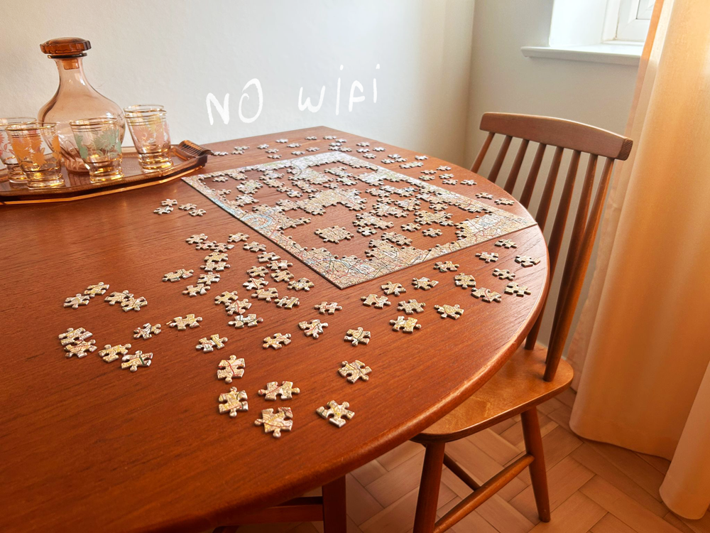 A wooden dining table with scattered puzzle pieces, a puzzle partially assembled, a glass carafe, glasses, and the words 'NO WIFI' written on the image.