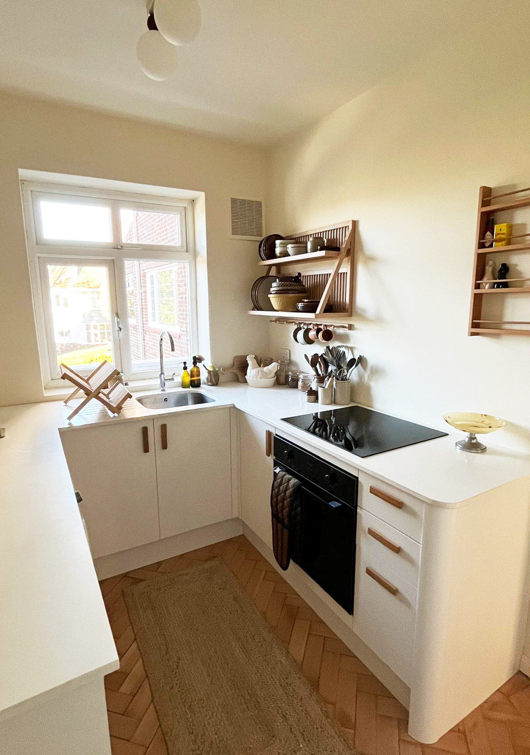 Cosy kitchen with white cabinets, wooden shelves holding dishes, a black oven, and a window letting in natural light.