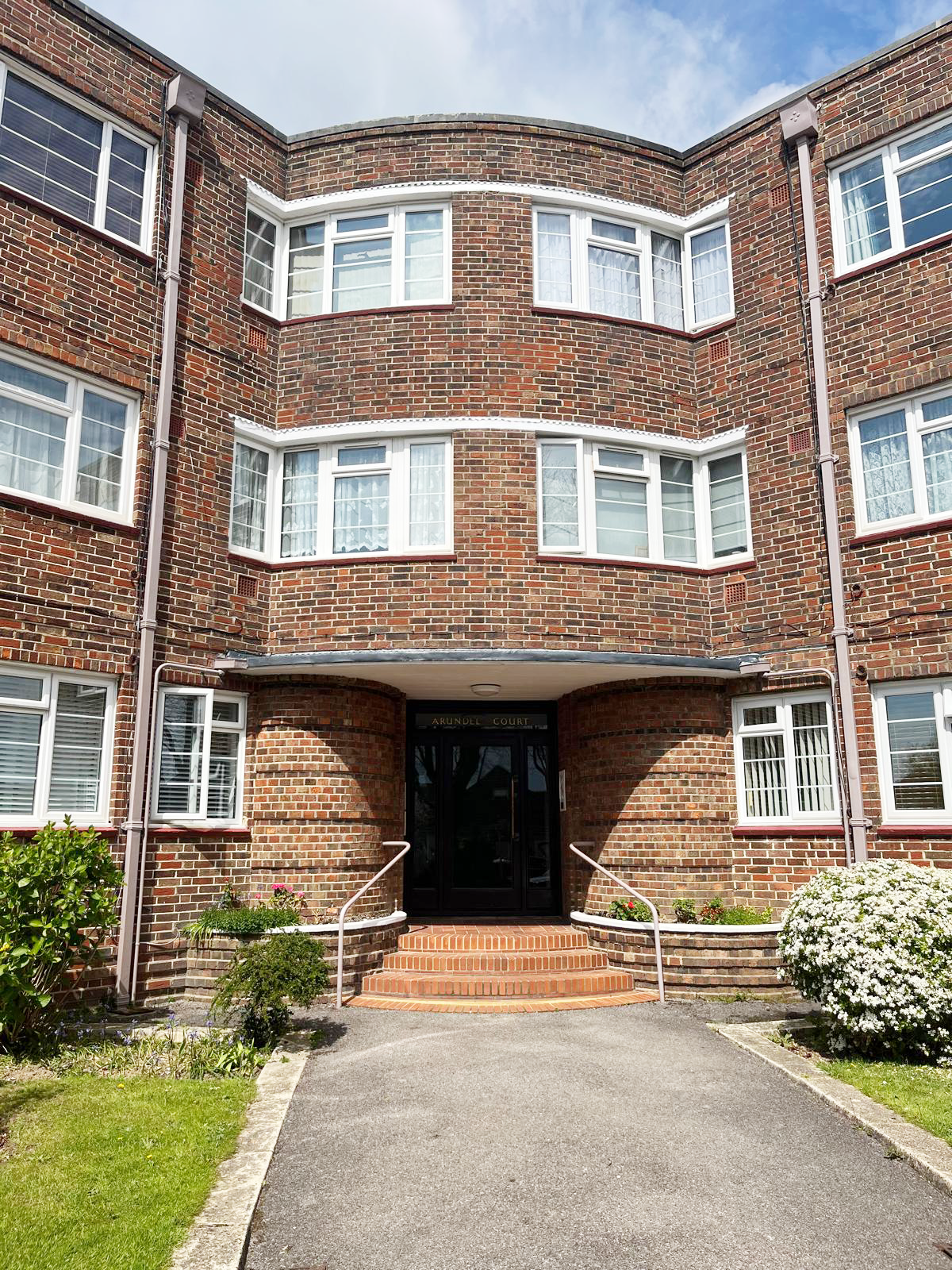 Brick apartment building with multiple white-framed windows and a black front door, set in a landscaped area with plants and a curved pavement.