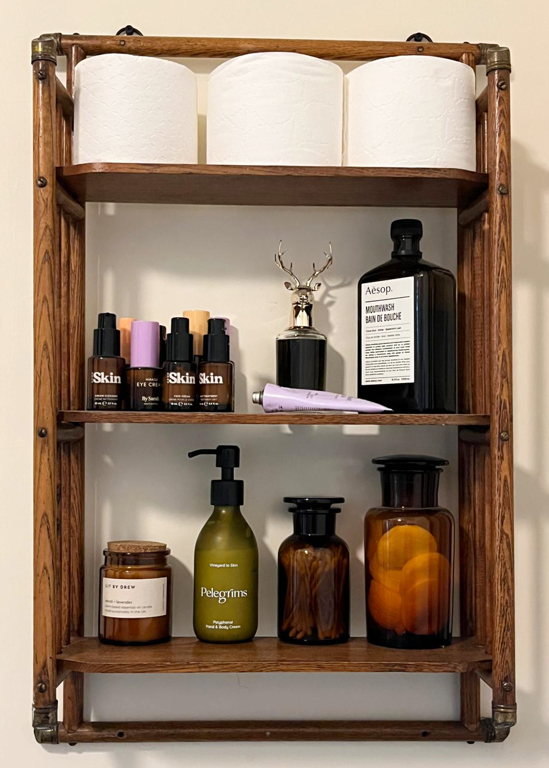 Wooden shelf with tissues on the top, skincare products, a small deer figurine, Aesop mouthwash, hand and body cream, and bottles on the lower shelves.
