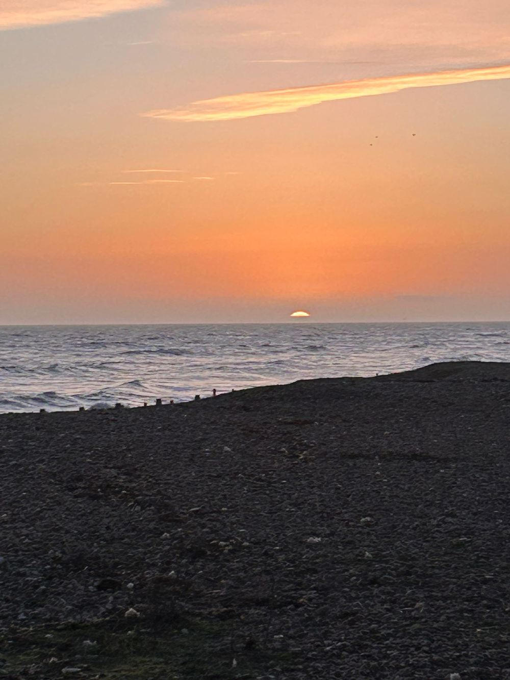 Sunset over the sea with a partly cloudy sky and a rocky beach foreground.