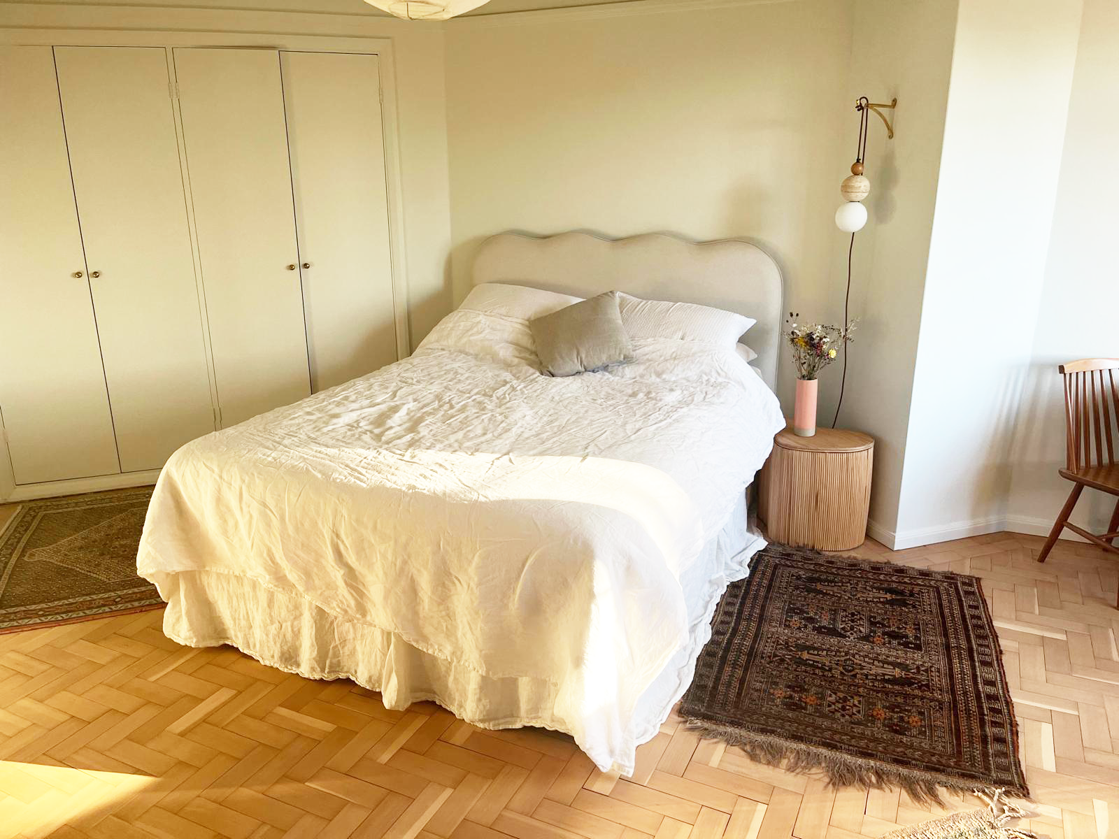 A bedroom with a white bed, a wooden nightstand with a vase of flowers, a wall-mounted lamp, a large rug, and a closed closet with four doors.