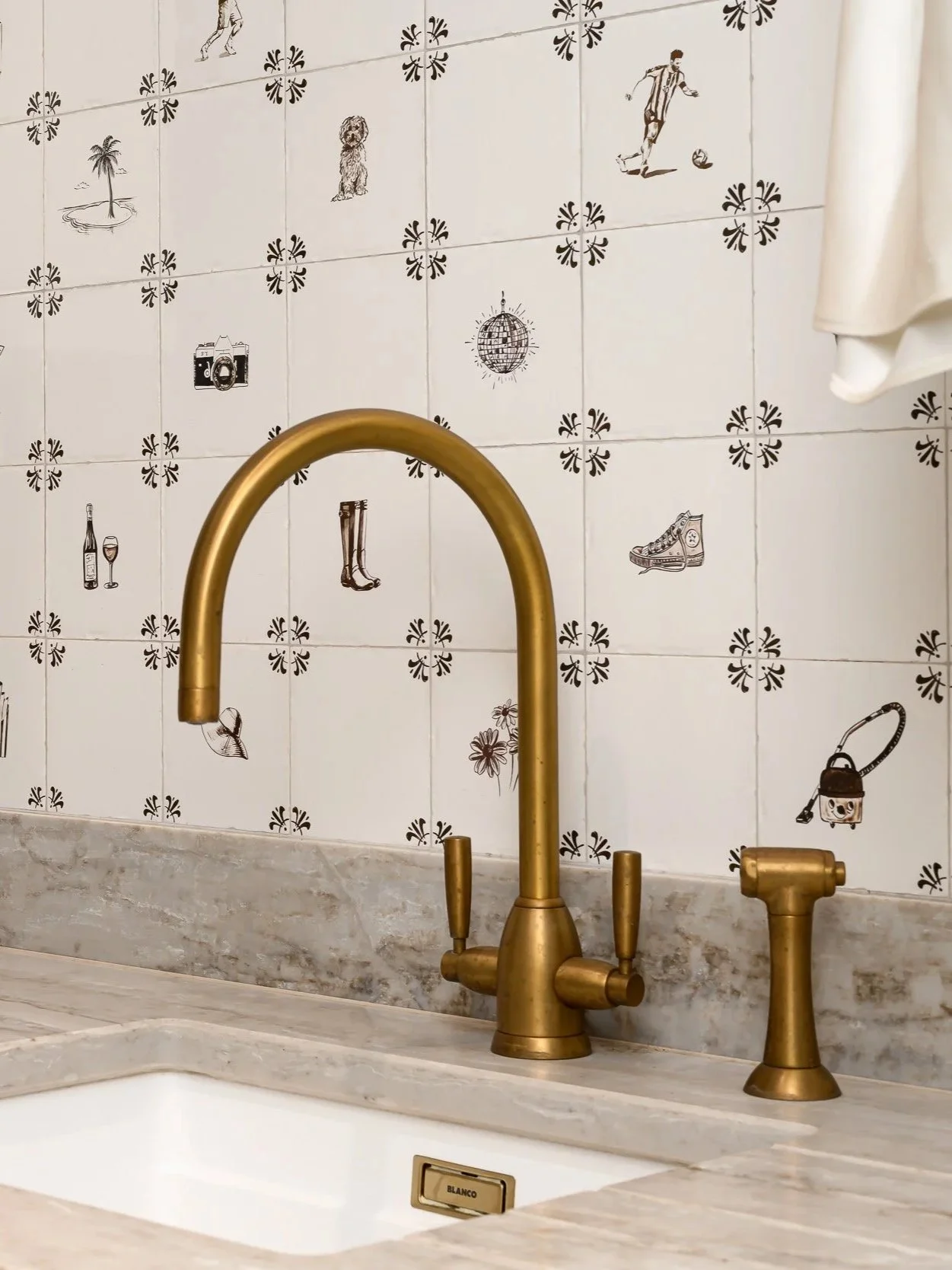 Kitchen sink with a gold-colored faucet and handle, and a marble countertop. Tiled wall behind the sink features black and white illustrations of various objects, including a palm tree island, a dog, a person playing soccer, a disco ball, a camera, a wine bottle with a glass, rubber boots, a sneaker, a vacuum cleaner, and some flowers.