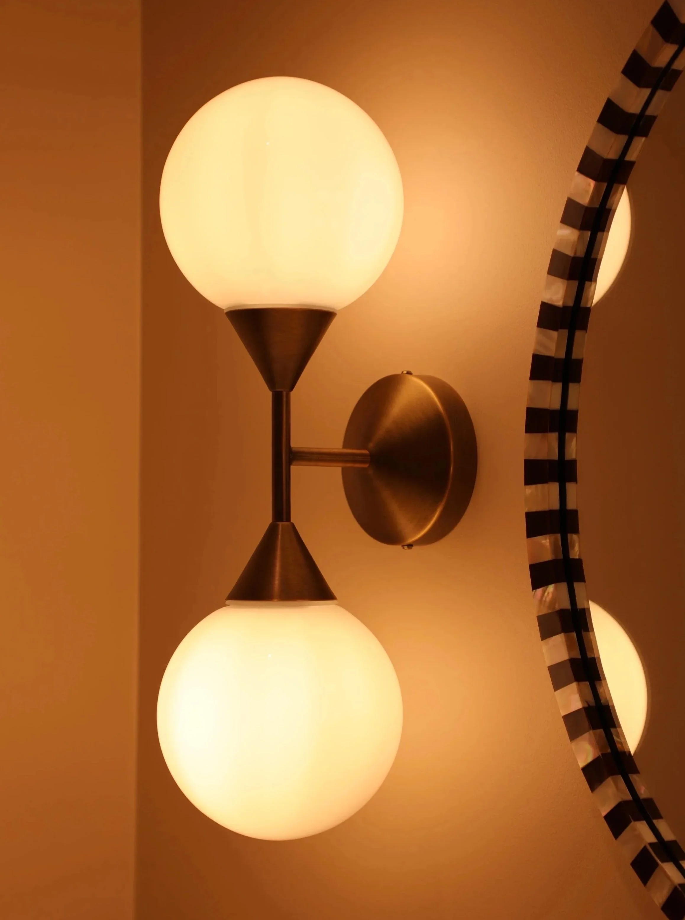 A wall-mounted light fixture with three large frosted glass globes and a brass finish, emitting warm light in a room with beige walls and a black-and-white patterned curtain.