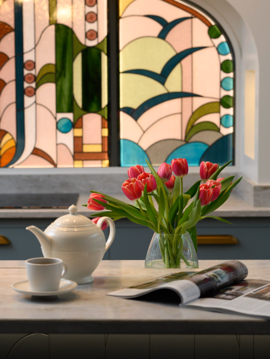 A kitchen counter with a white teapot, a cup of coffee, a magazine, and a glass vase with pink tulips, with a stained glass window featuring abstract shapes and colors in the background.