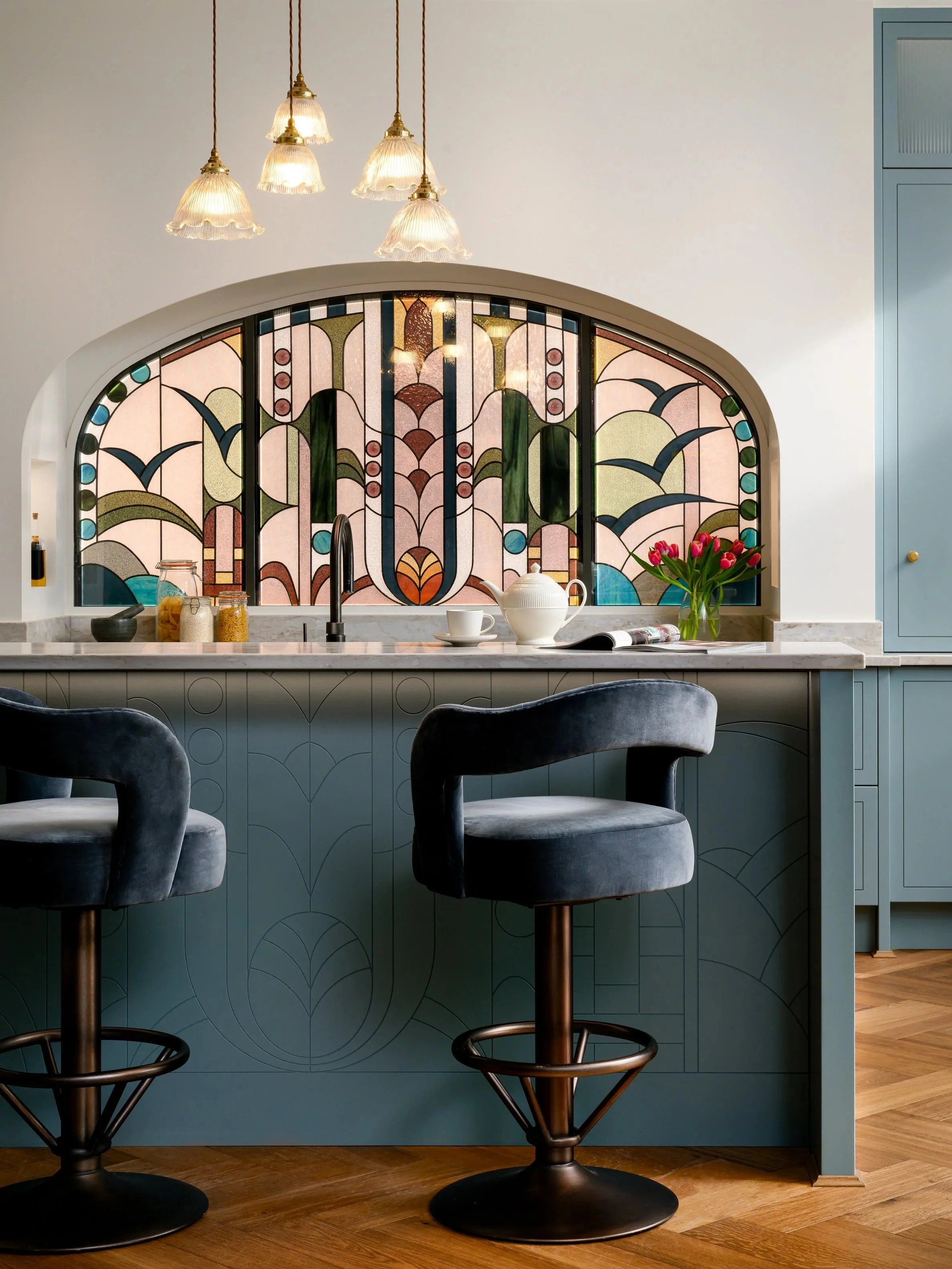 Kitchen with a blue island, stained glass window, black bar stools, and a teapot with pink tulips on the counter.