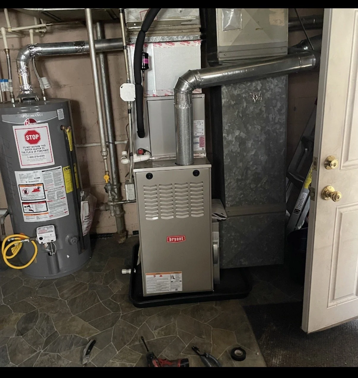 A utility room containing a modern furnace and a water heater, with various pipes and ductwork connected to the heating and water systems. The floor is tiled, and there are tools and a tape measure on the floor.