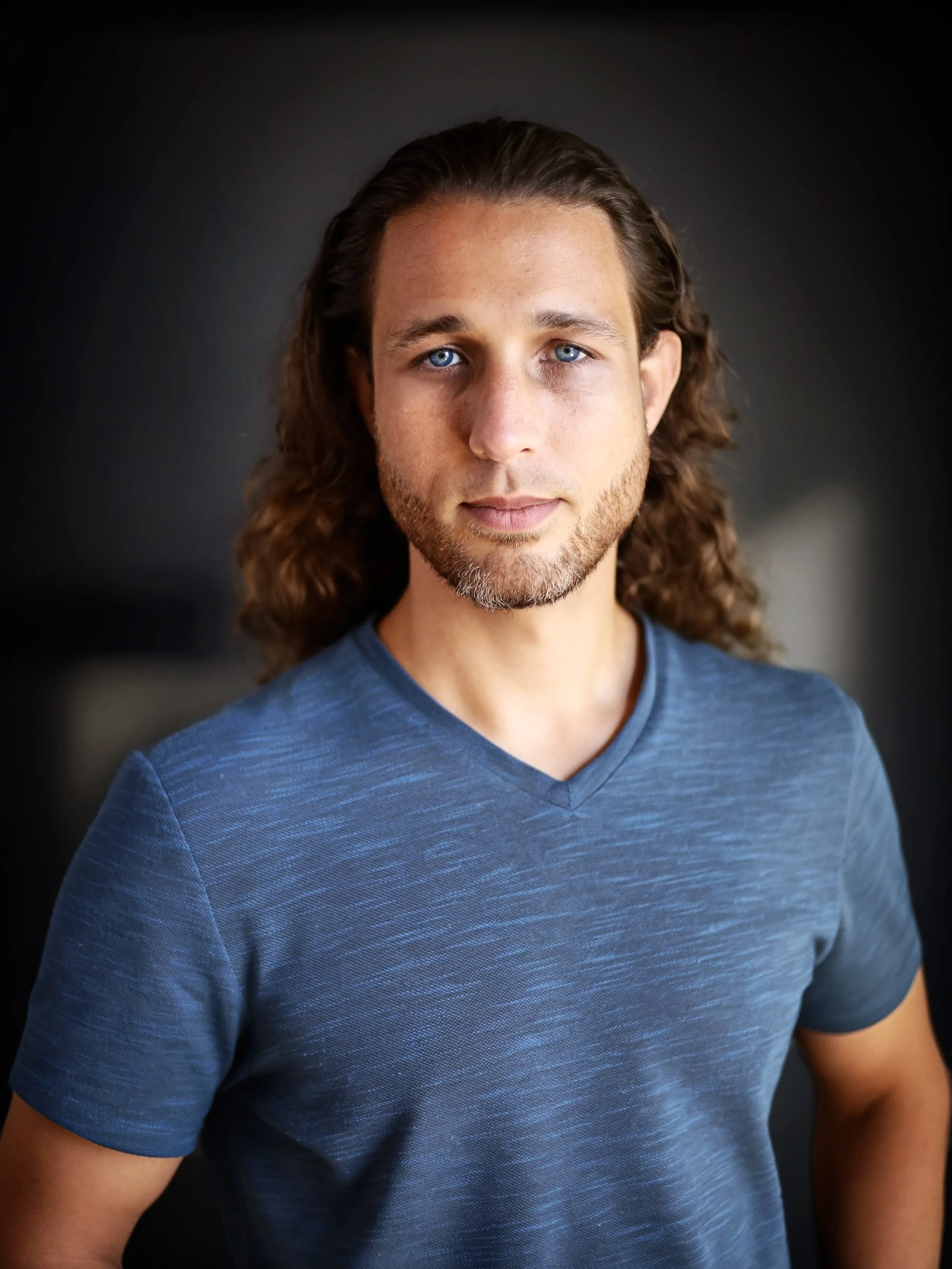 Close-up portrait of a young man with blue eyes, long brown hair, a light beard, wearing a blue V-neck shirt, against a dark background.