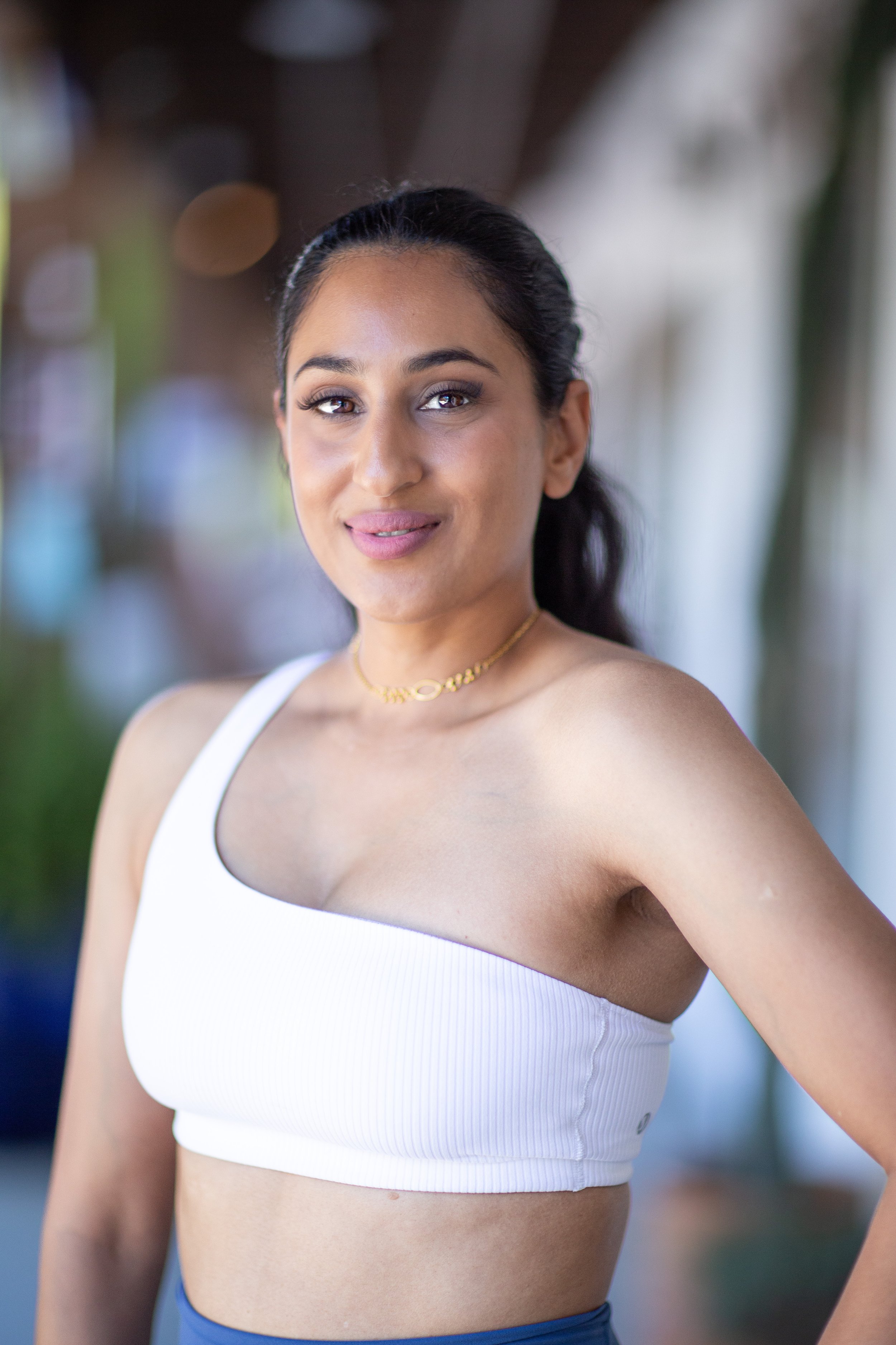 A woman in a white tank top and blue bottoms standing outdoors with a blurred background, smiling and looking at the camera.