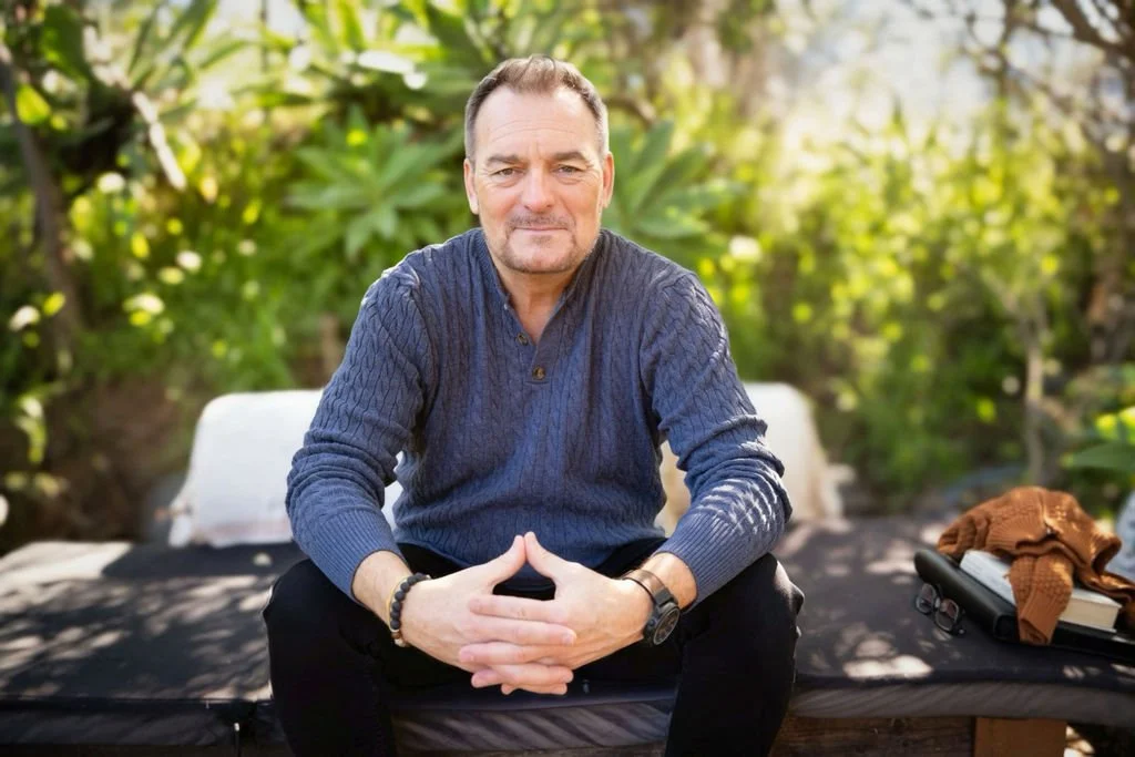 A middle-aged man with short dark hair and a beard, sitting outdoors on a black lounge chair, wearing a blue sweater, black pants, and a watch, with nature greenery in the background.