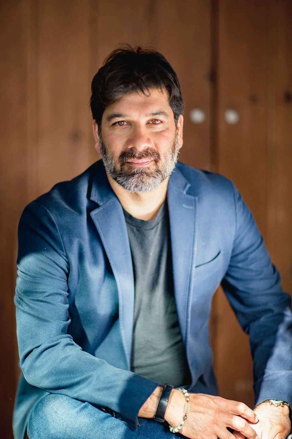 A man with dark hair and a beard, wearing a dark blazer over a t-shirt, sitting in front of a wooden background.