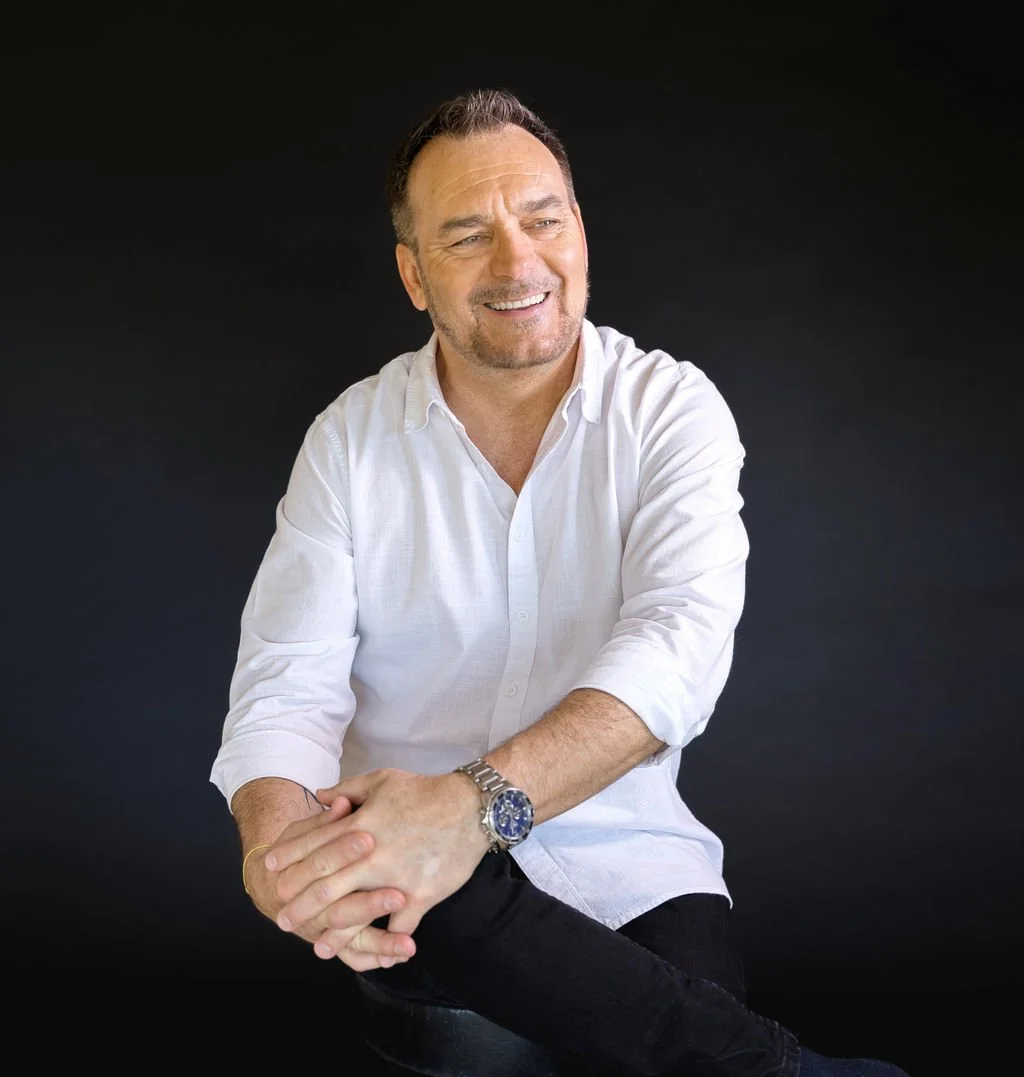 A smiling middle-aged man with short hair, wearing a white button-up shirt, black pants, a wristwatch, and a gold bracelet, sitting against a black background.