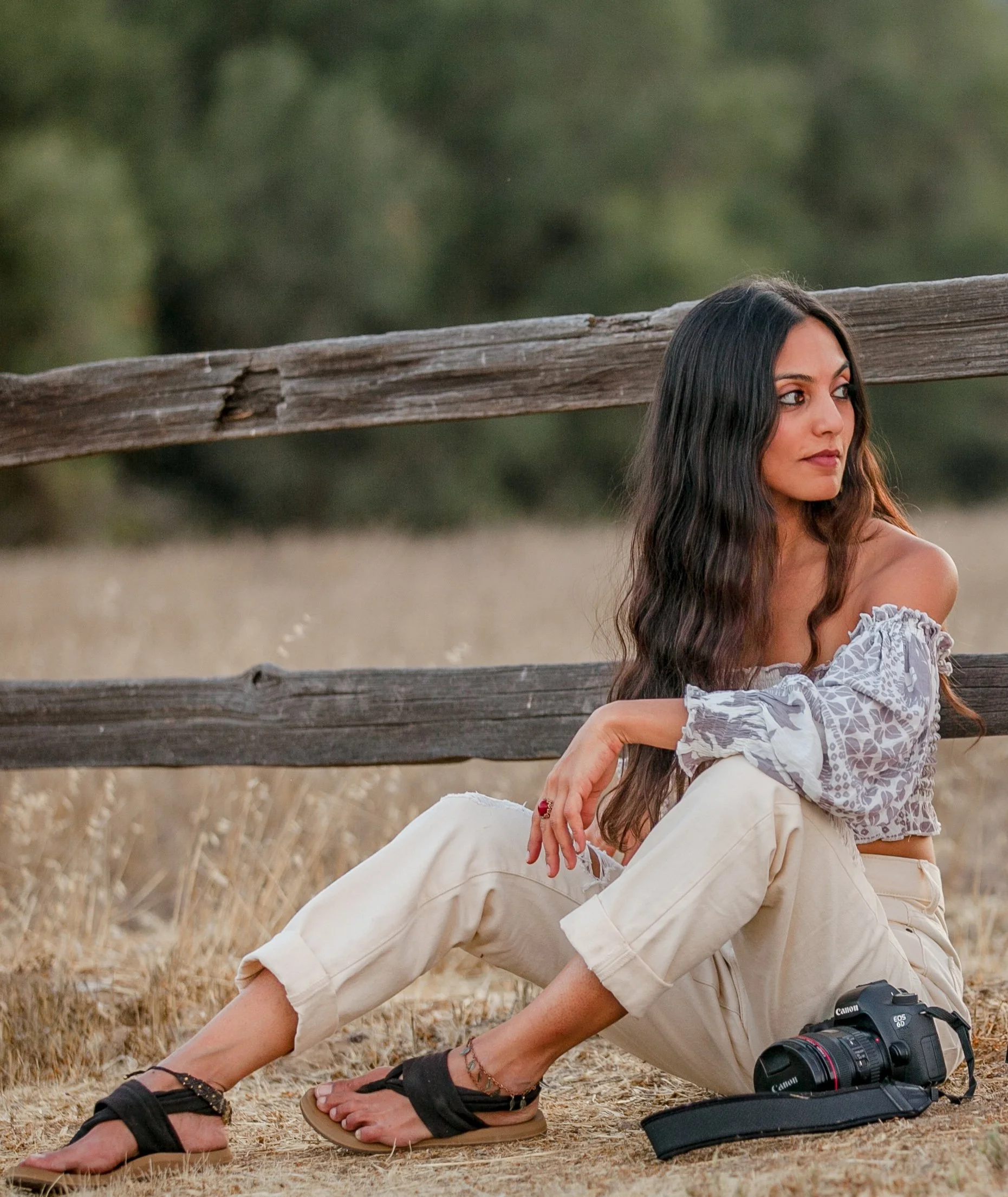 A woman sitting on dry grass near a wooden fence, wearing beige pants, a patterned off-shoulder top, and sandals, with a camera placed on the ground nearby.