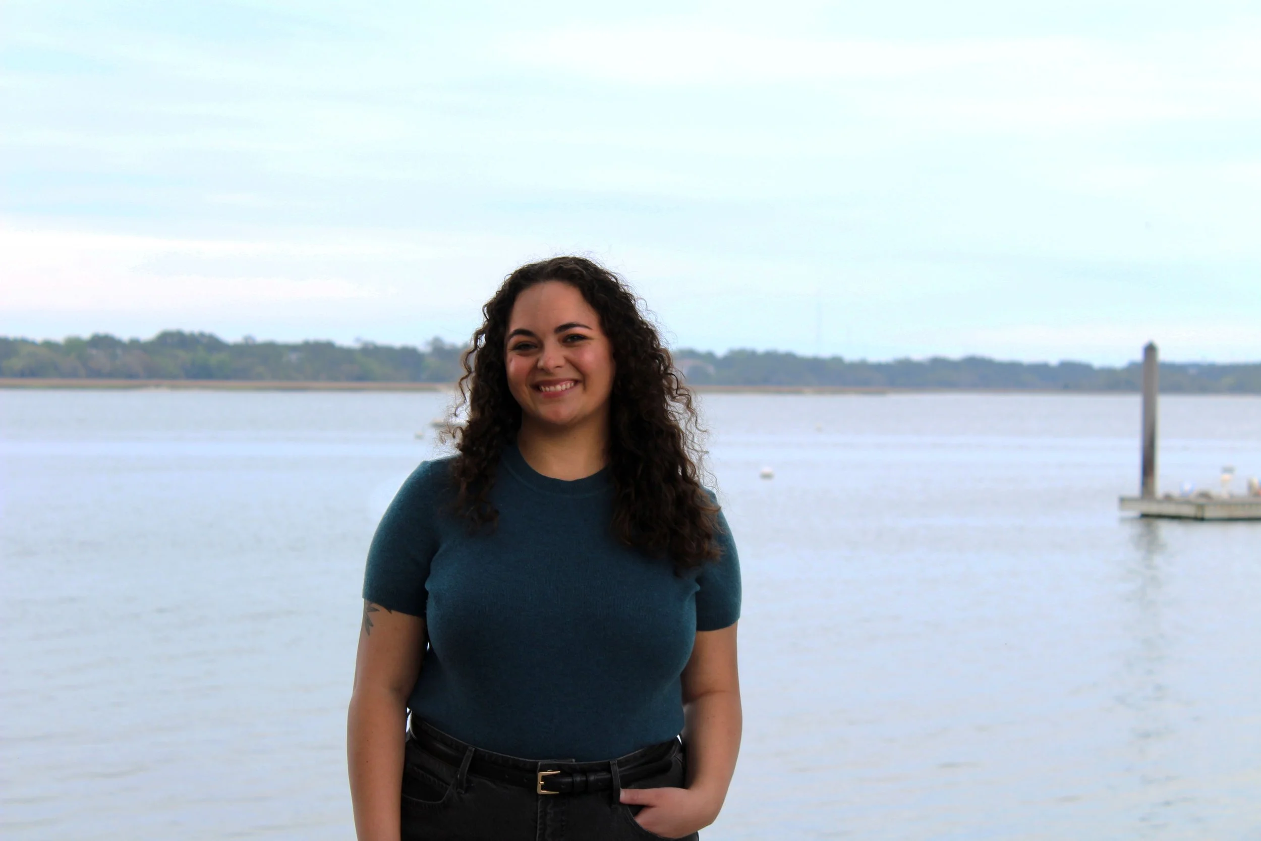 A woman with curly dark hair, wearing a teal t-shirt and black pants, smiling and standing by a body of water with a distant shoreline and cloudy sky in the background.