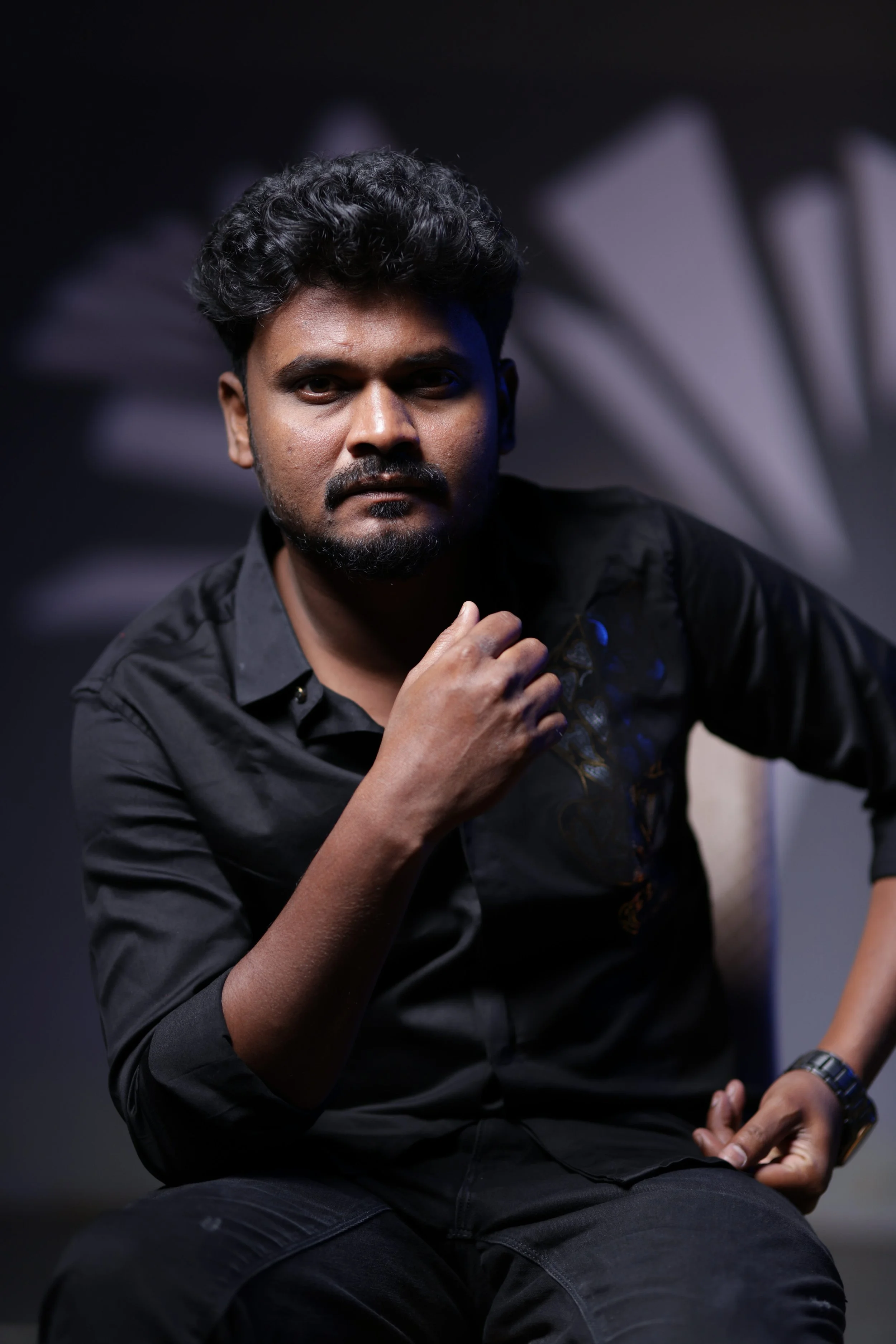 A man wearing a black shirt with an intricate design on the chest, sitting with his hand on his chin, in front of a dark background with blurred geometric shapes.