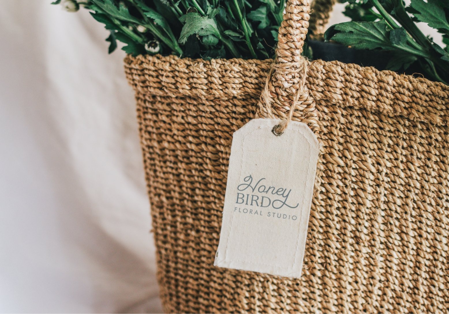 A woven basket filled with green plants. Attached to the basket is a cream-colored tag that reads 'Honey BIRDY FLORAL STUDIO'.