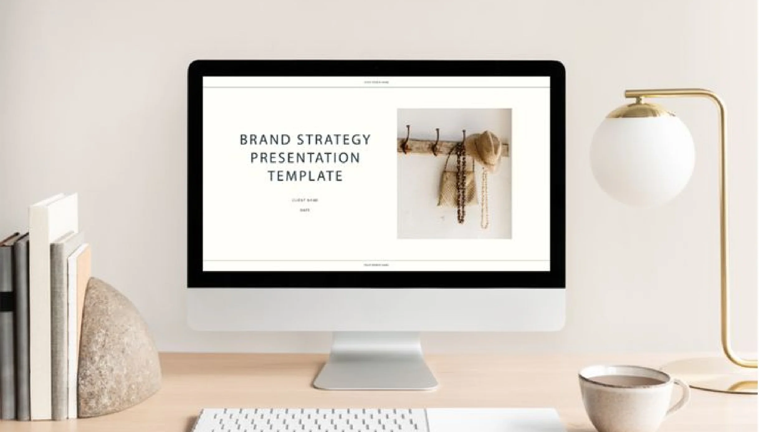 Computer monitor displays a presentation slide titled 'Brand Strategy Presentation Template' with a small photo of hooks and hanging items on a wall, in a minimalist workspace with books, a rock, a cup, and a lamp.