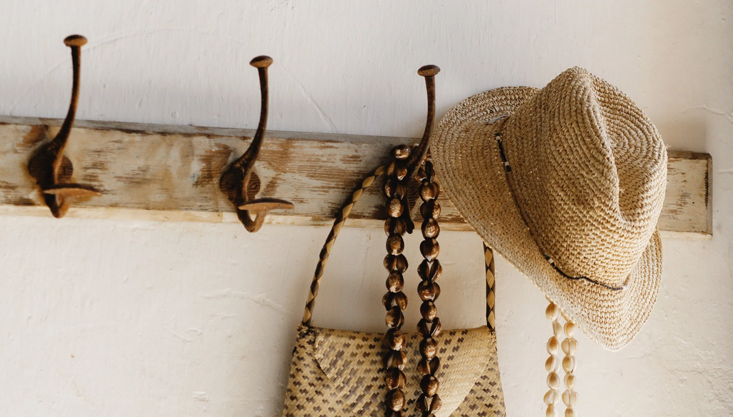 A rustic wooden coat rack with three metal hooks, holding a straw hat and hanging necklaces made of shells and beads, against an off-white wall.