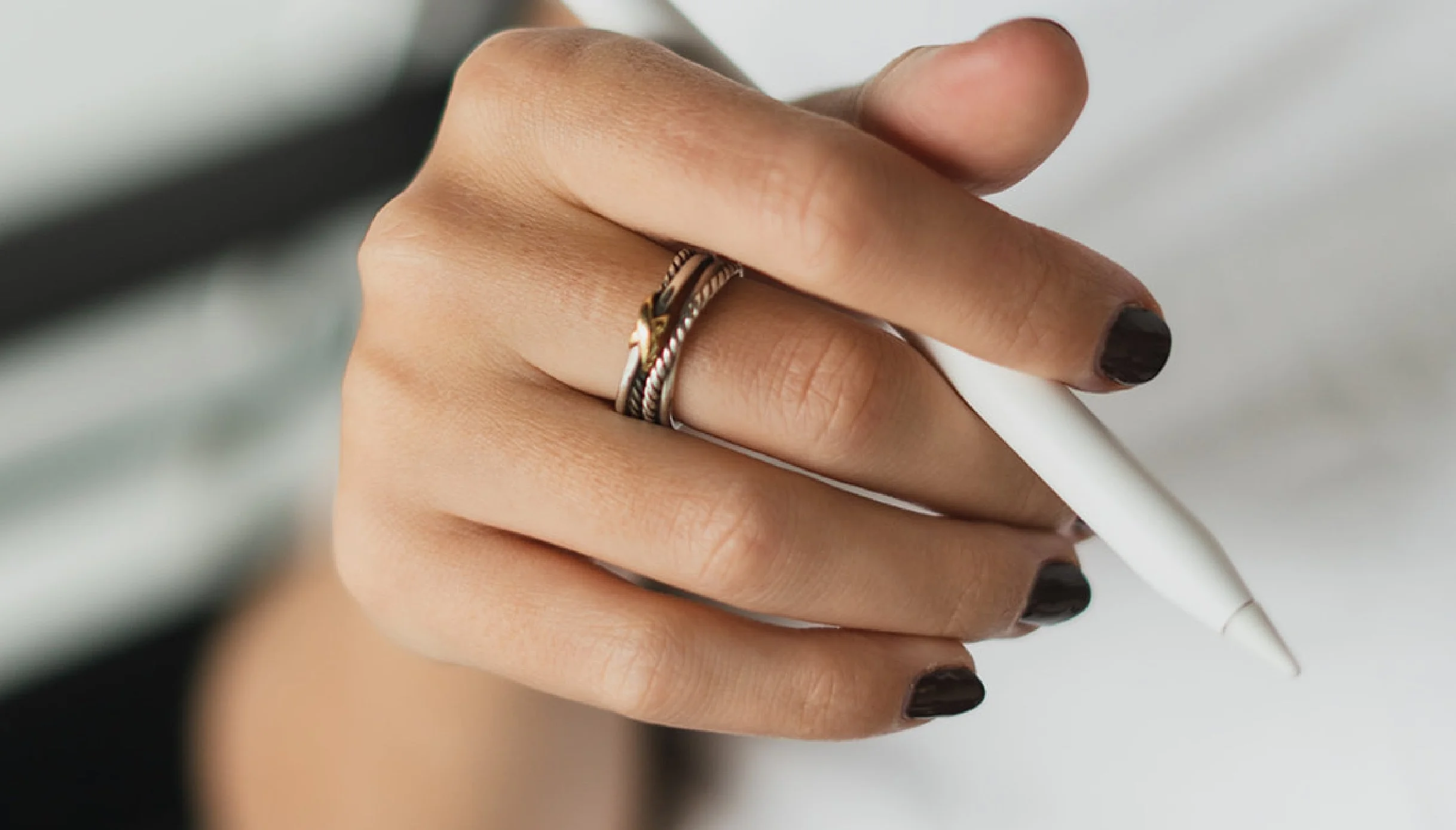 Close-up of a hand holding a white pen, with dark nail polish and wearing two rings on the ring finger, against a blurred background.