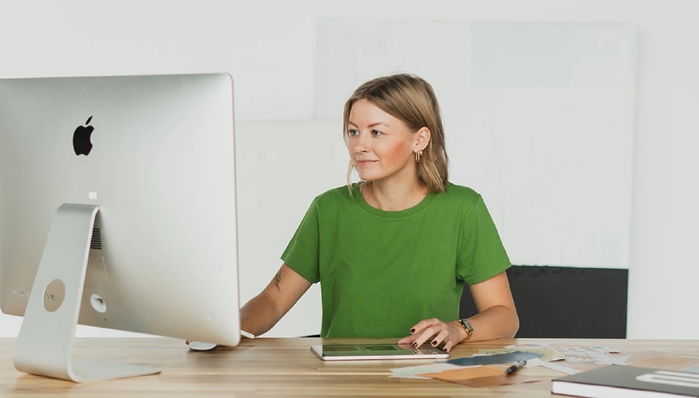 Woman in green shirt working at a desk with a large Apple computer, color samples, and a tablet.