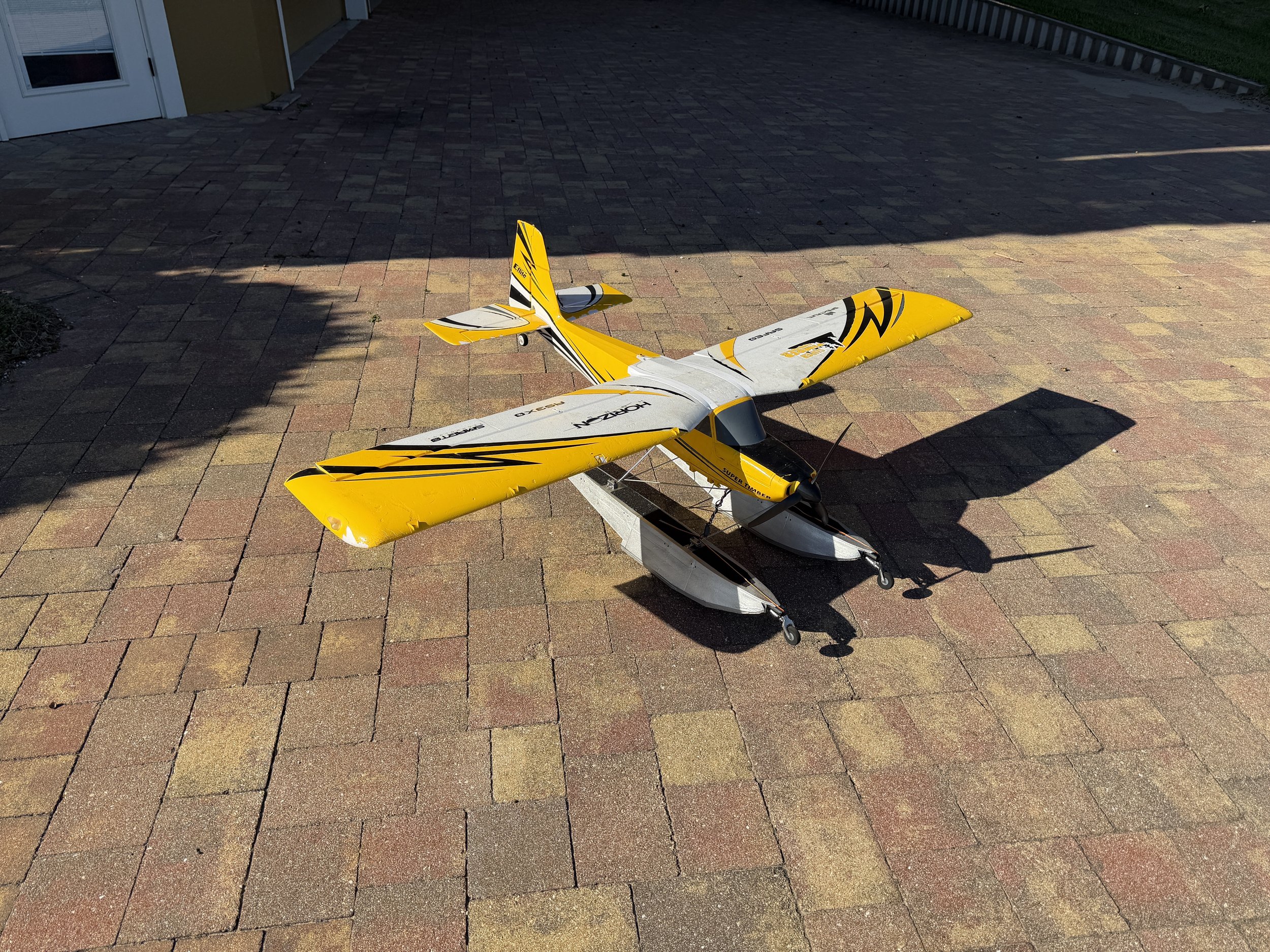 Yellow and white remote-controlled model airplane on a brick patio with houses and shadows.