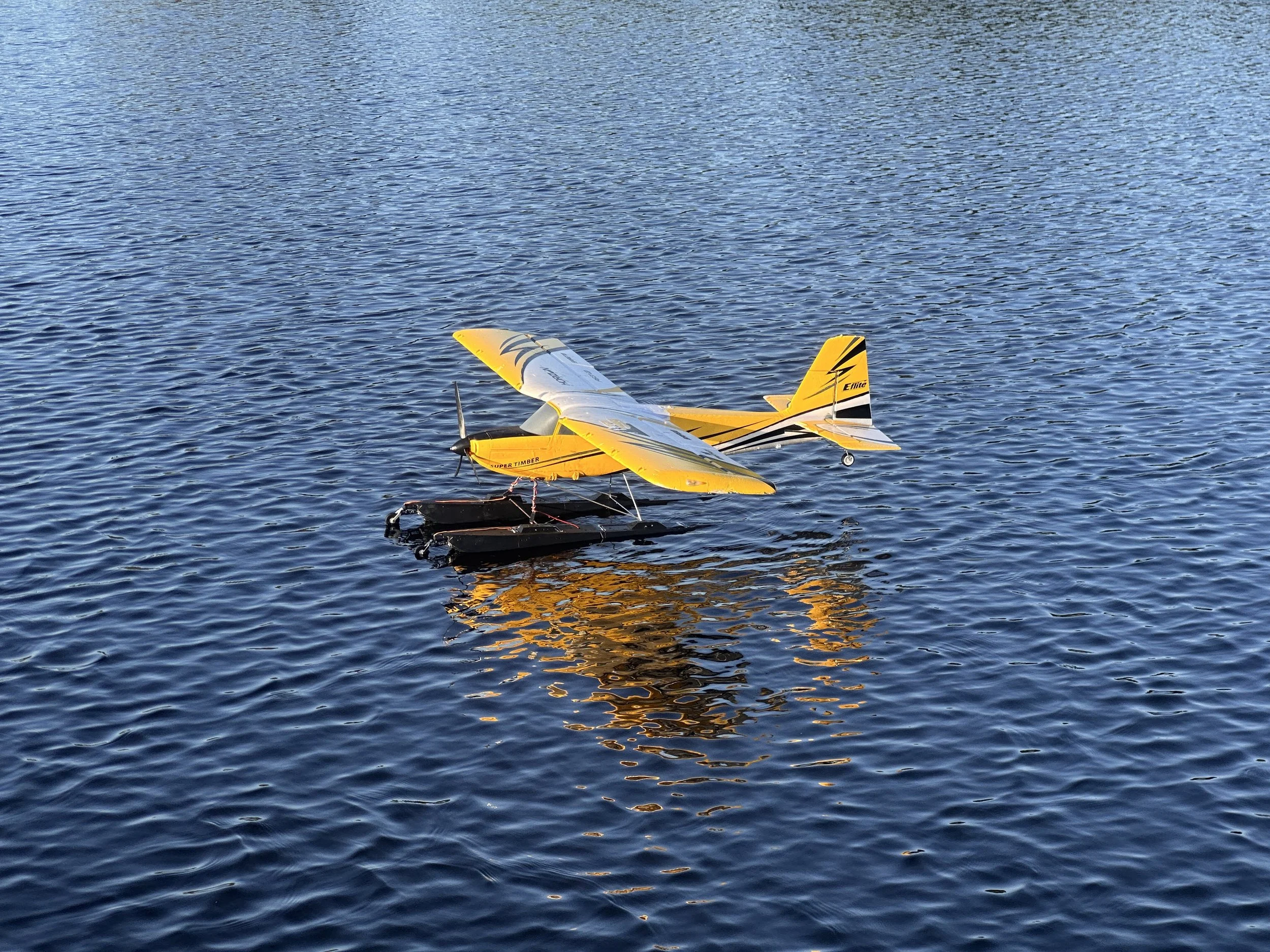 A yellow radio-controlled seaplane floating on calm water with reflections.