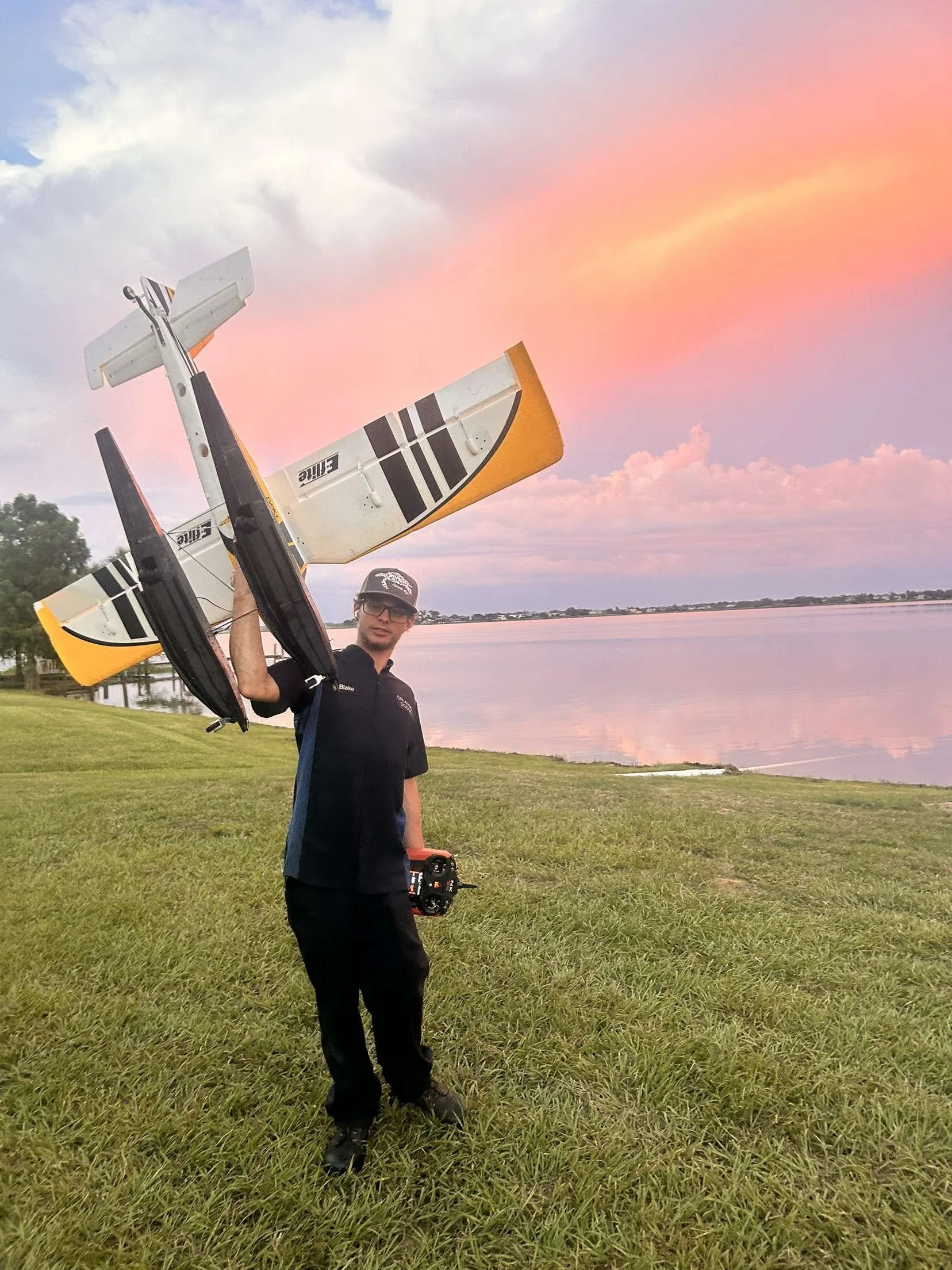 A man standing on grass near water holding a remote control and a large radio-controlled airplane during sunset, with a pink and purple sky.