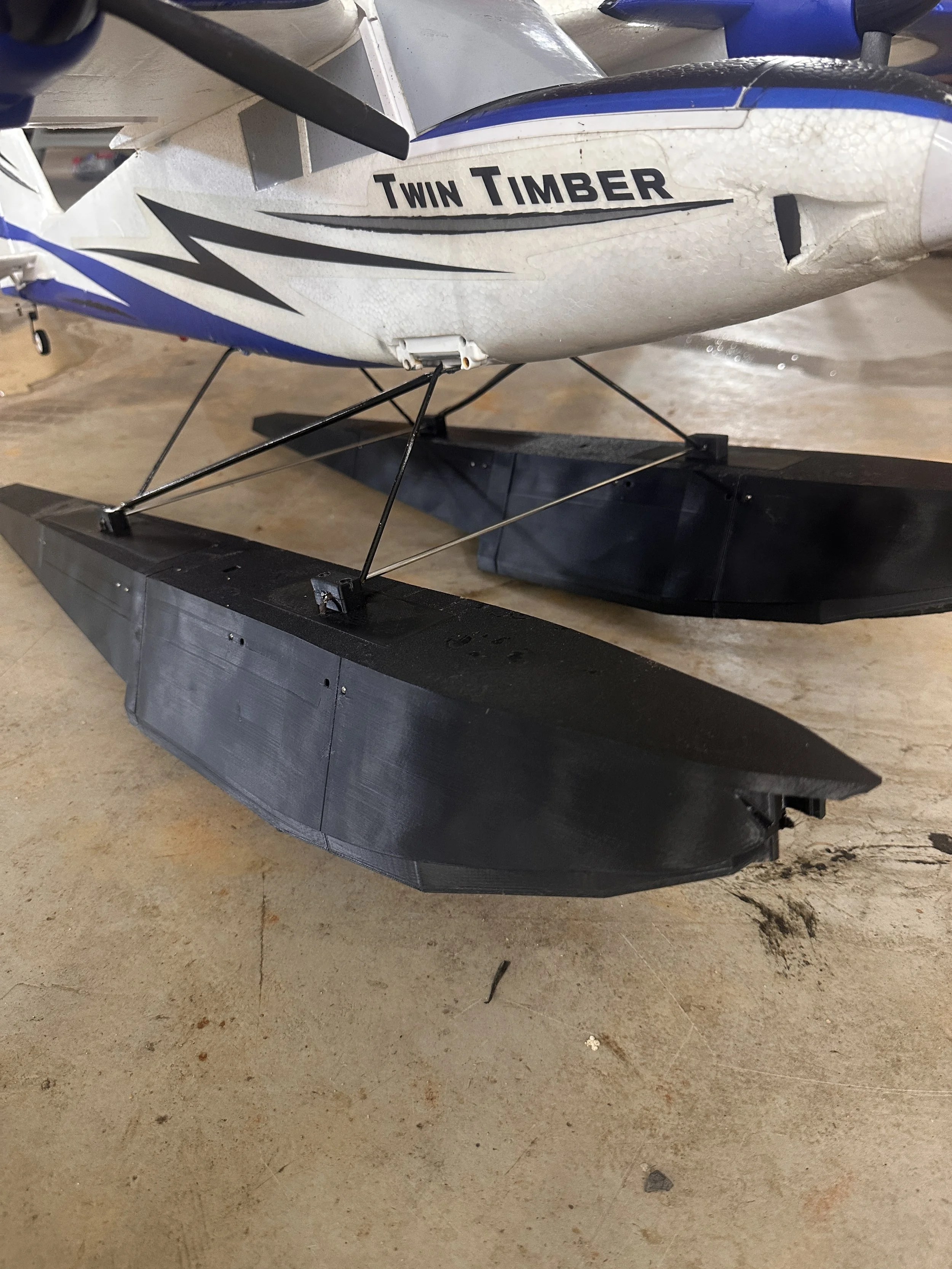 Close-up of a remote-controlled model airplane with the text "Twin Timber" on the side, mounted on a black plastic or composite ski for gliding on snow or ice, on a concrete floor.
