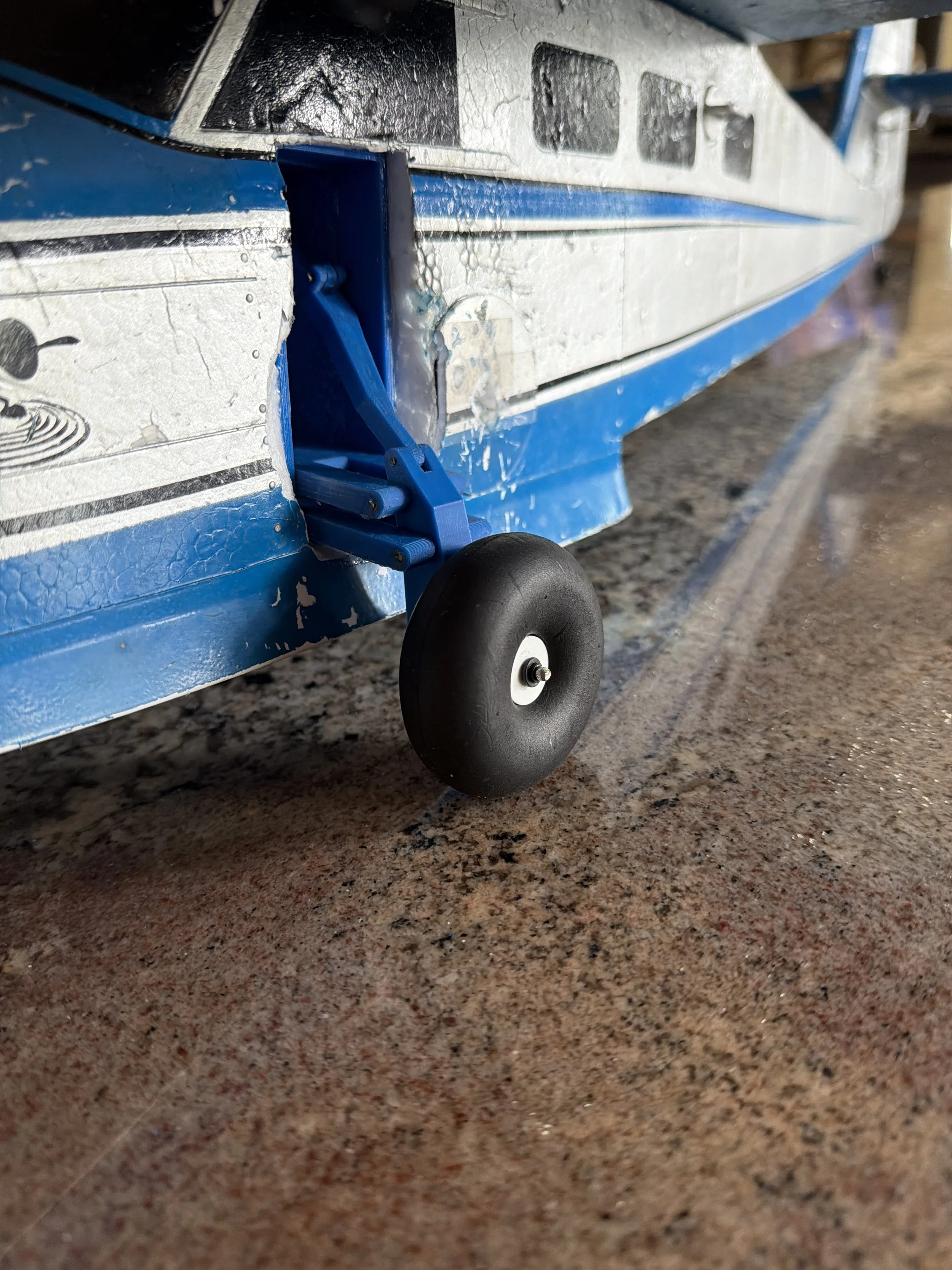 Close-up of a small wheel attached to a blue and white model boat on a textured surface.