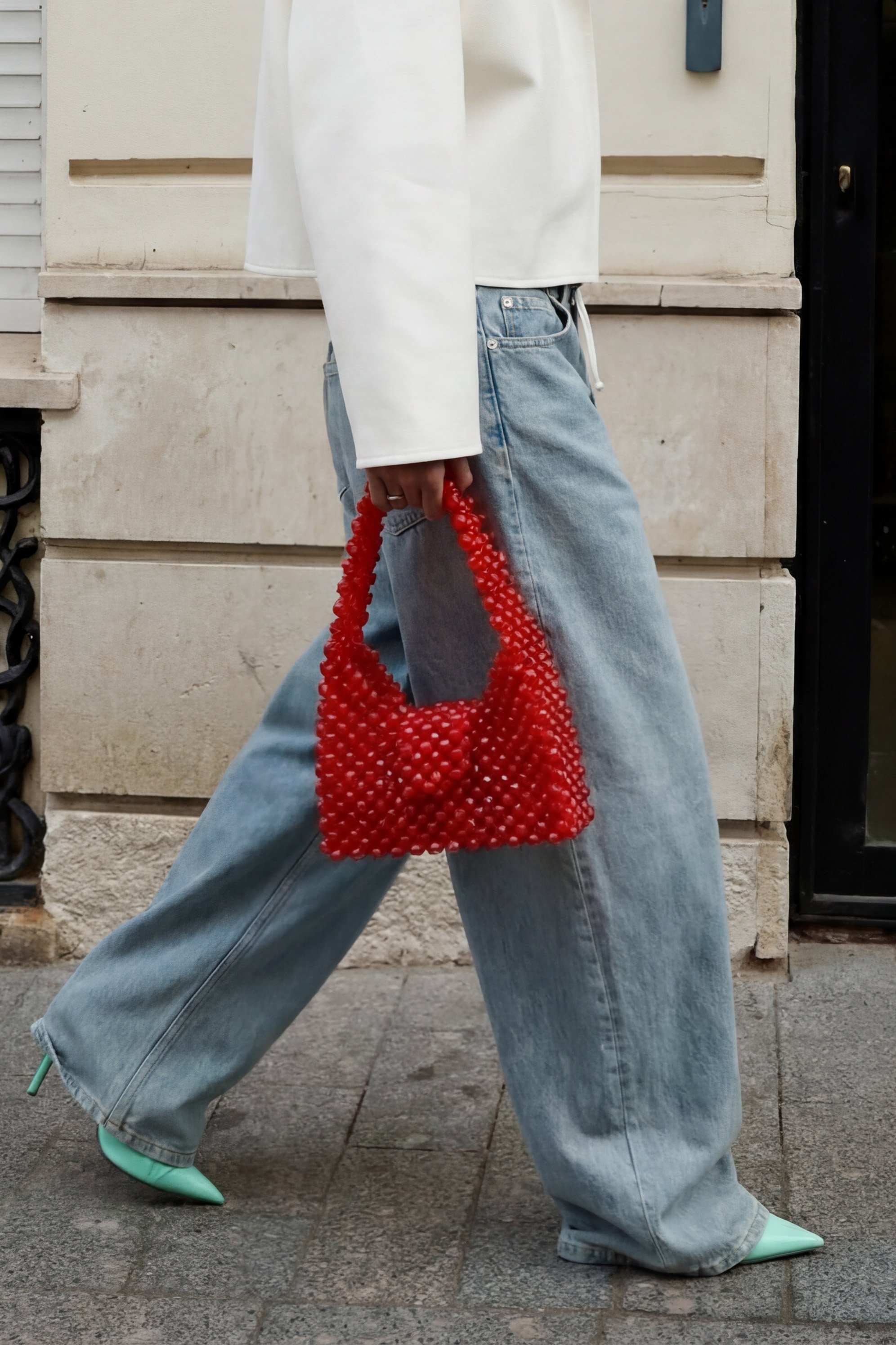 Person walking on sidewalk wearing light blue wide-leg jeans, mint green pointed shoes, a white top, and holding a red beaded handbag.