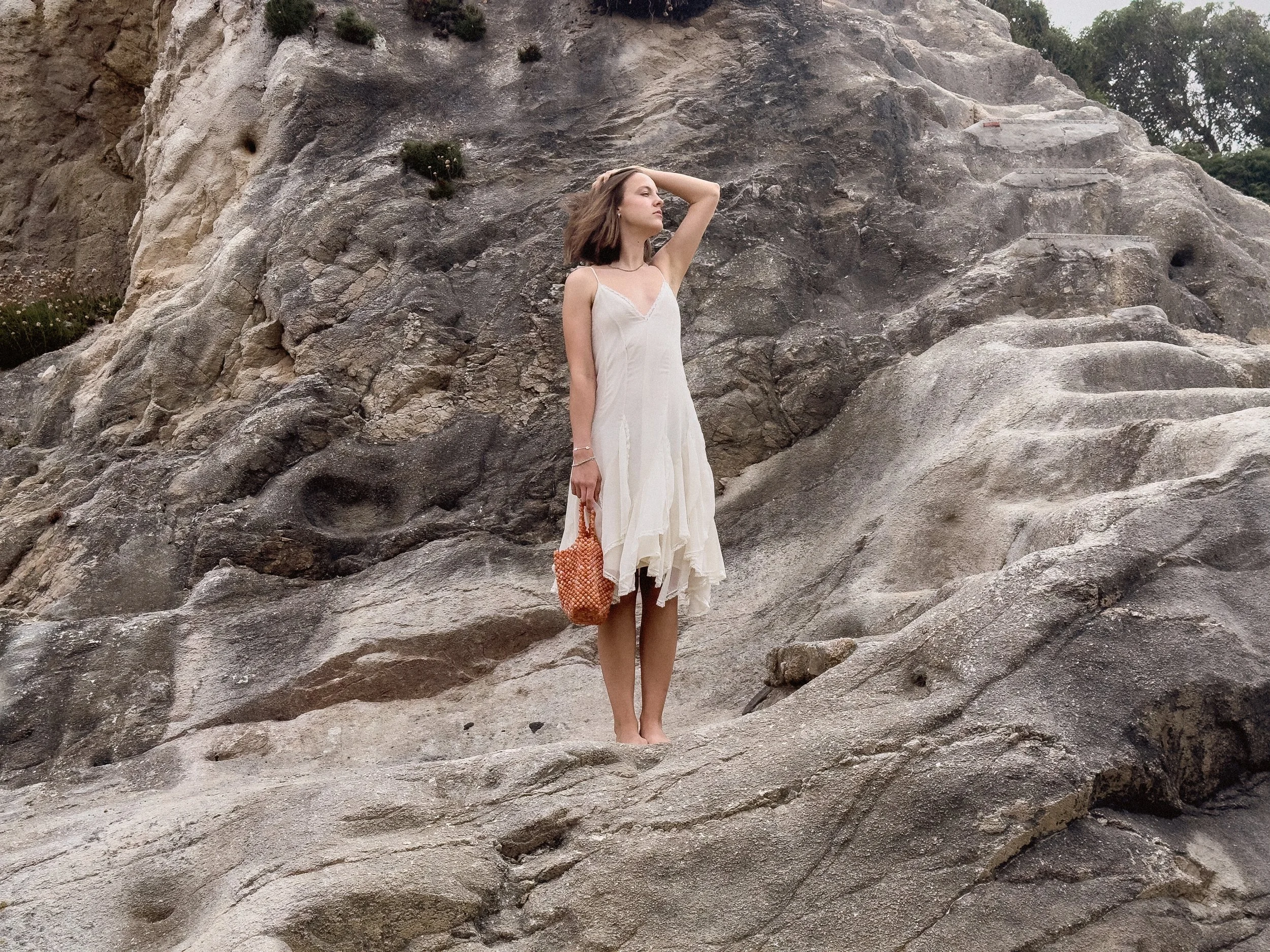 A woman in a white dress pauses on a rocky beach, holding an orange bag, with a large rock formation and greenery in the background.