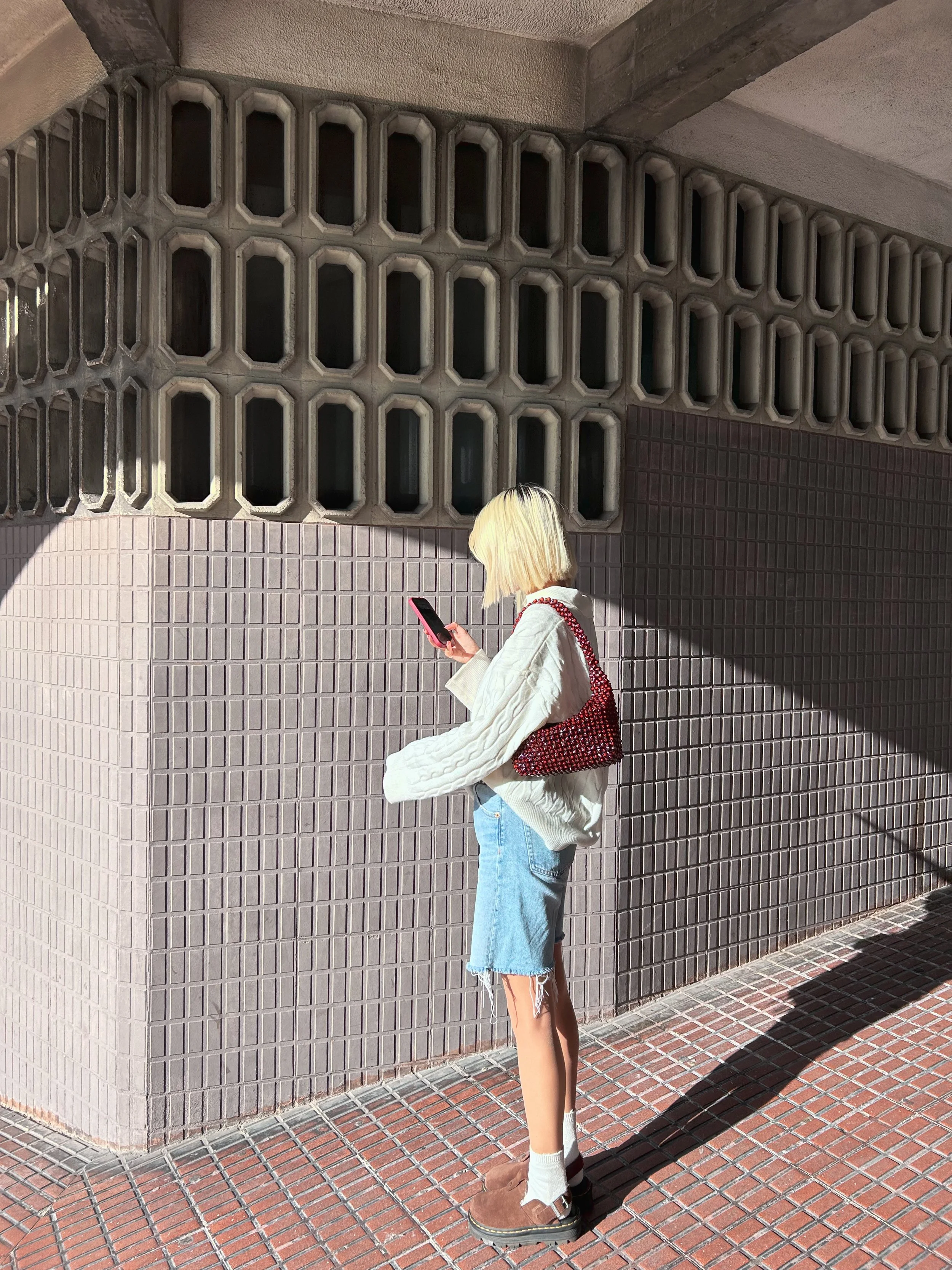 A young woman with blonde hair, wearing a white oversized sweater, denim shorts, white socks, and brown shoes, stands on a brick sidewalk and looks at her phone against a patterned concrete wall with sunlight casting a shadow.