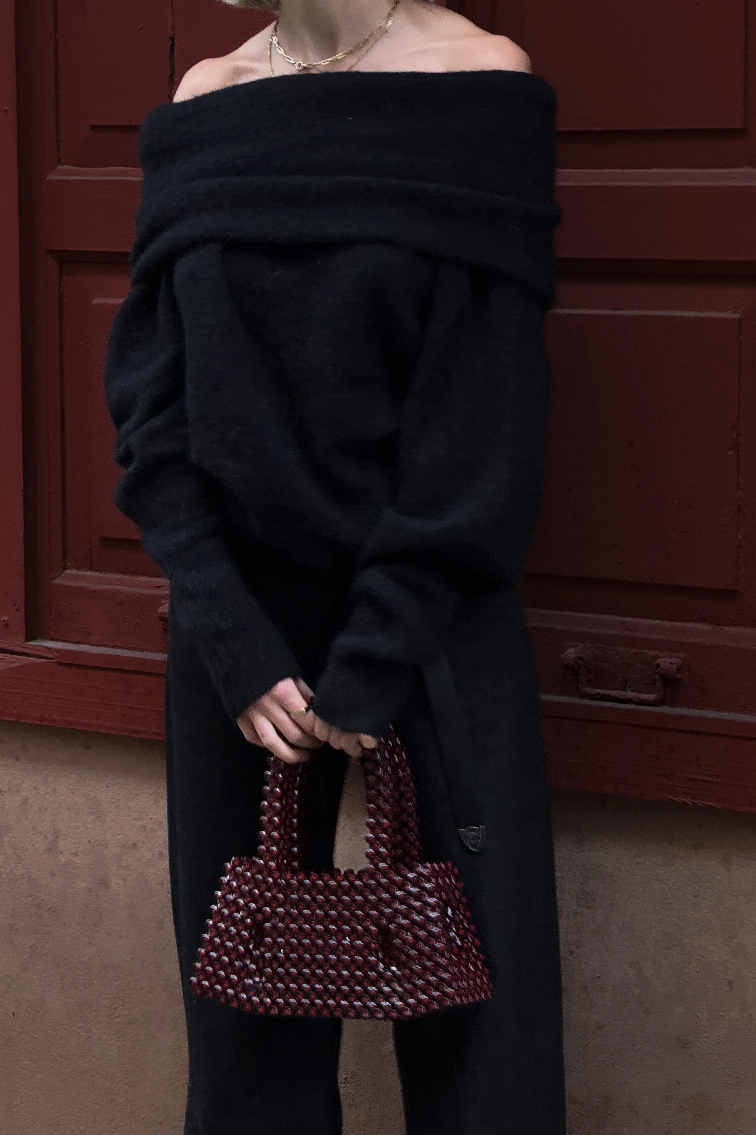 Person dressed in a black off-shoulder sweater and black pants, holding a red and black patterned handbag, standing in front of a wooden red door.