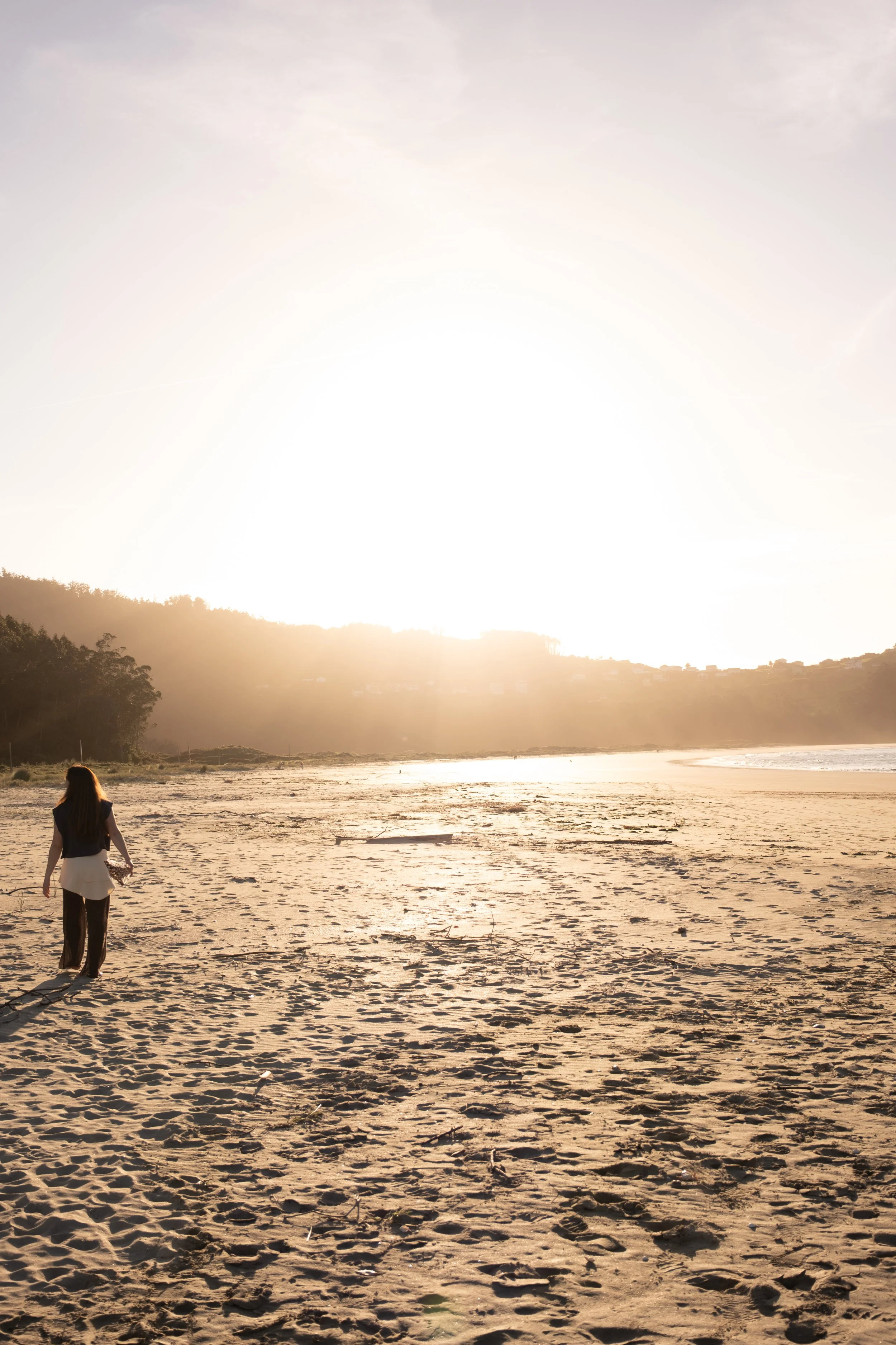 A woman walking on a sandy beach at sunset or sunrise with hills and trees in the background.