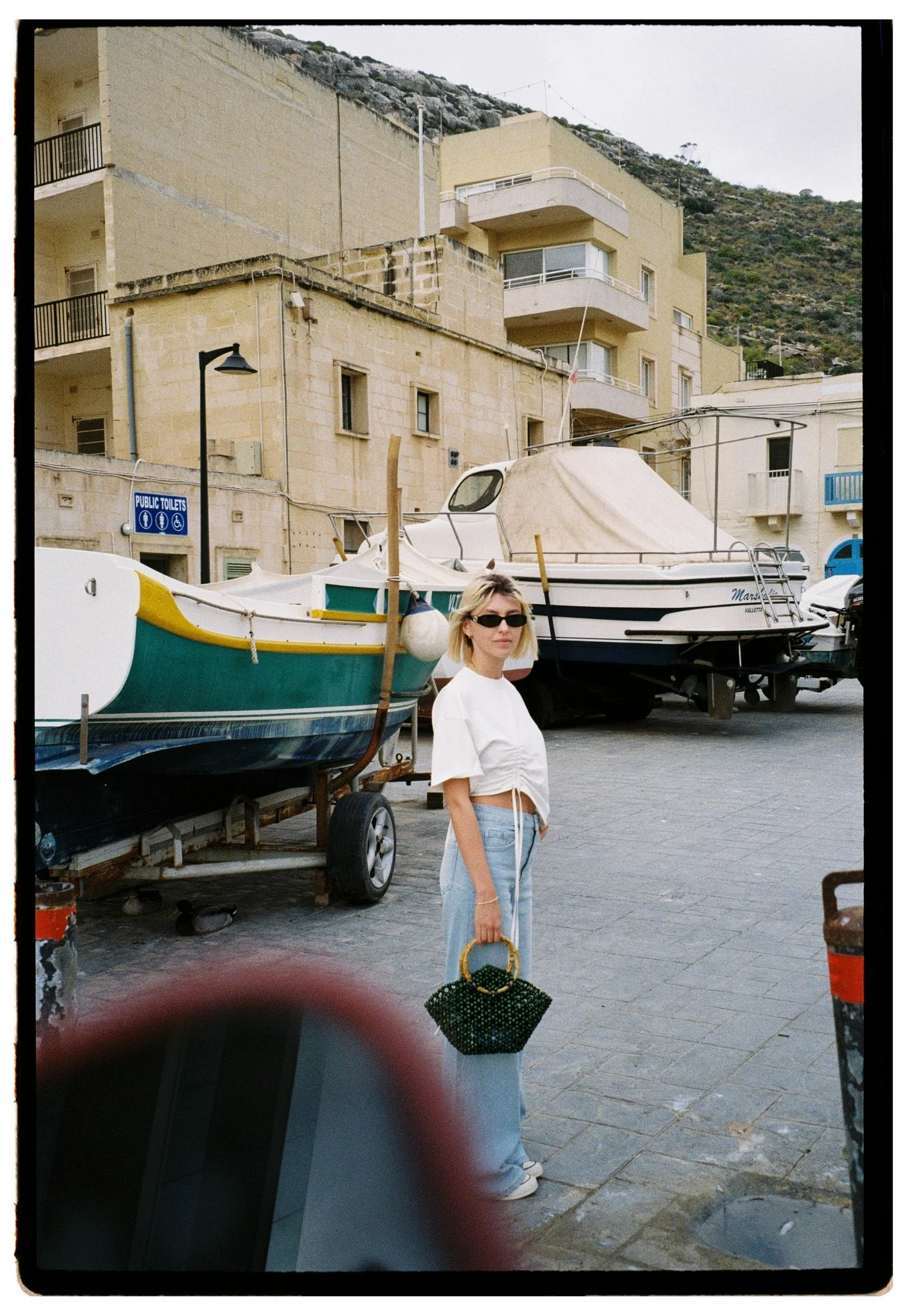 A woman in a white crop top and light blue jeans standing on a paved street next to boats on trailers, with buildings and a hillside in the background.
