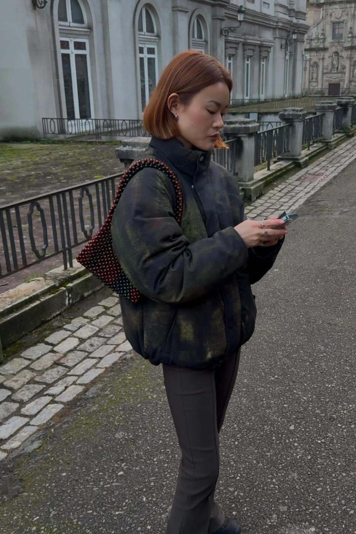 A woman with shoulder-length reddish-brown hair, dressed in a dark jacket and trousers, stands on a cobblestone sidewalk, looking at her phone. She carries a beaded bag on her shoulder, and the background features an ornate, historic building with large windows and stone railings.