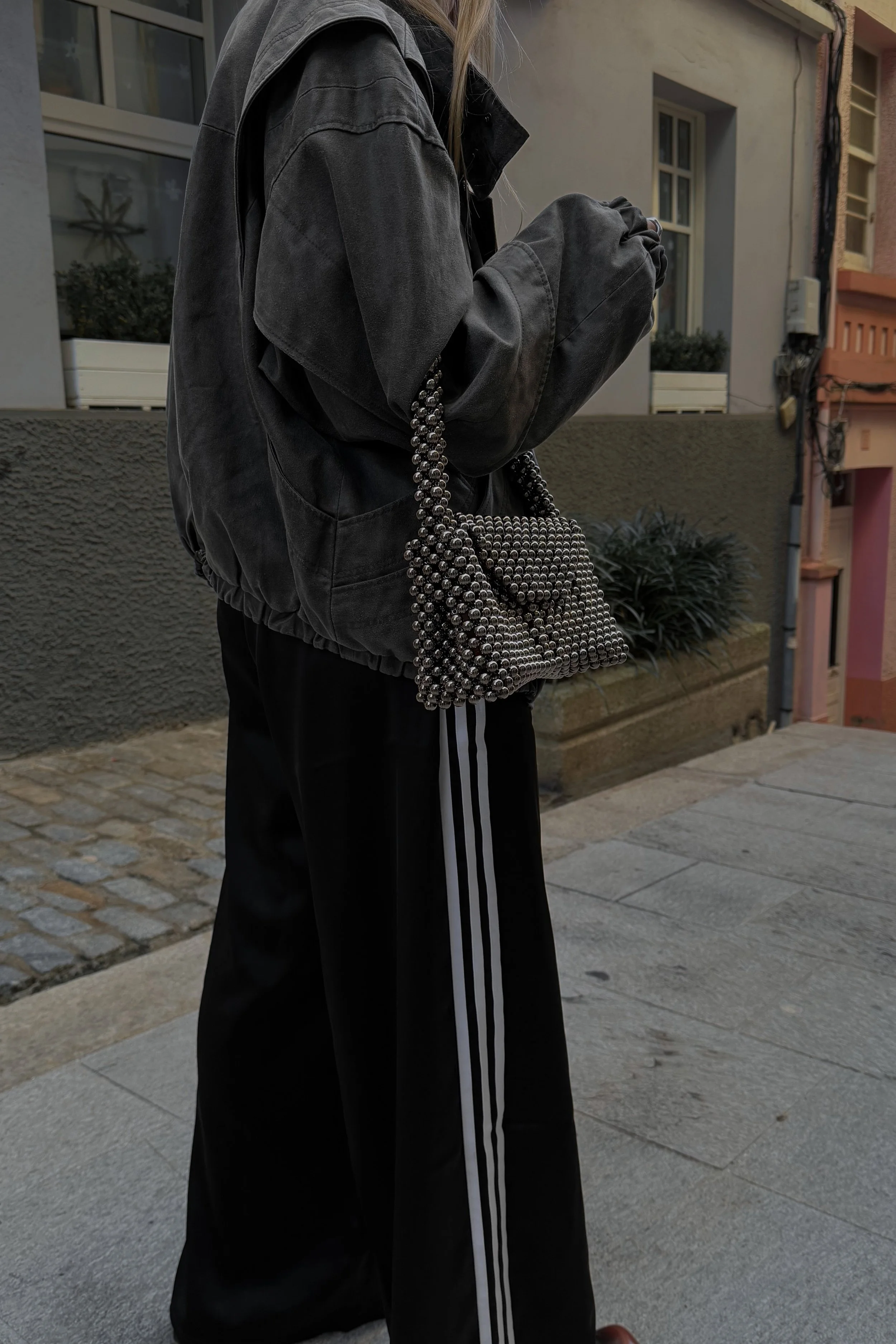 Person dressed in black casual clothing, carrying a handbag made of silver metal balls, standing outdoors near a building with windows and plants.