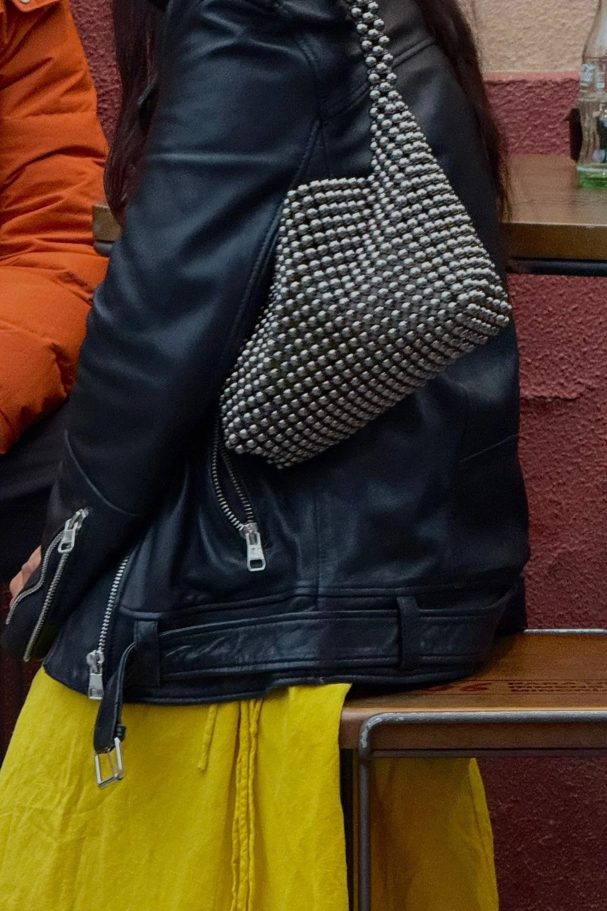 Person wearing a black leather jacket with silver zippers, yellow pants, and carrying a metallic beaded bag, sitting on a wooden bench against a red textured wall.